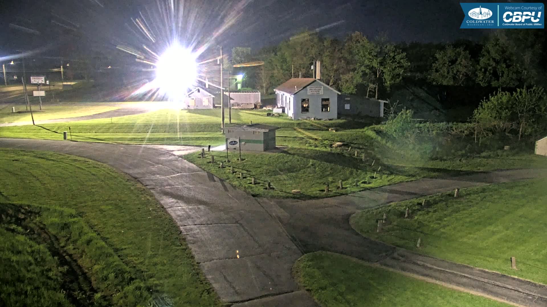 A nighttime view of a paved road intersecting with a grassy area, featuring several small buildings and trees under bright artificial light.