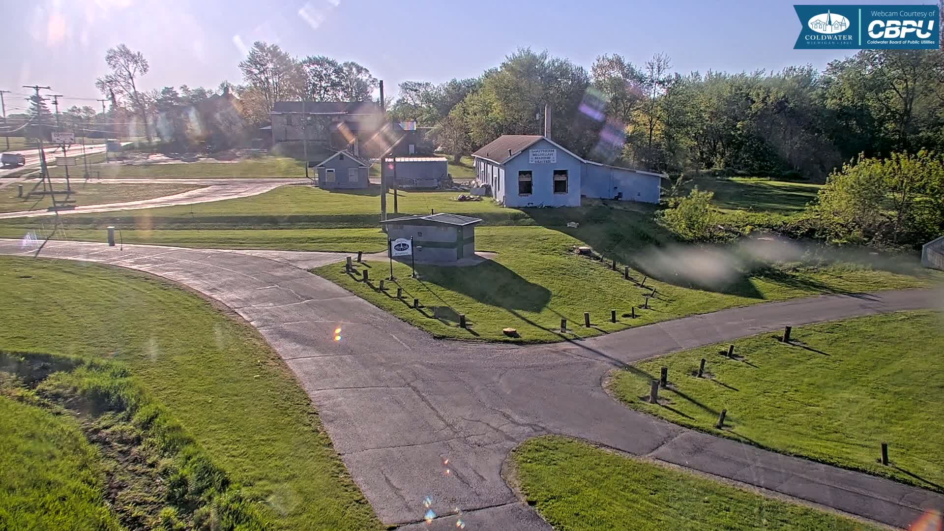 A sunny day reveals a paved pathway system intersecting near several small buildings and a grassy area, with trees and a road visible in the background.