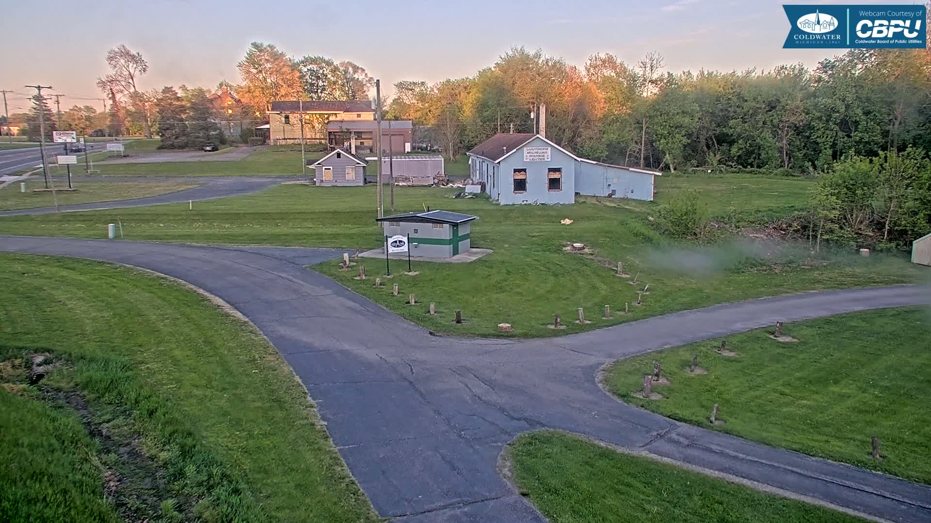 Under a clear twilight sky, a paved roadway curves through a grassy area with several small buildings and trees in the background.