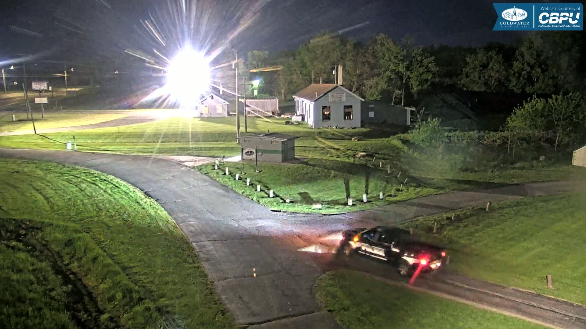 A dark-colored pickup truck drives on a paved road at night, past a grassy area with several small buildings, under bright artificial lights.