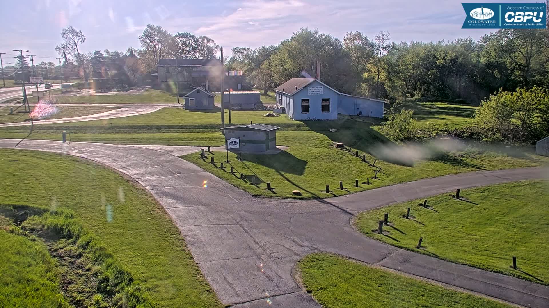 A paved path intersects near a small green building in a grassy area with several other buildings and trees under a mostly sunny sky.