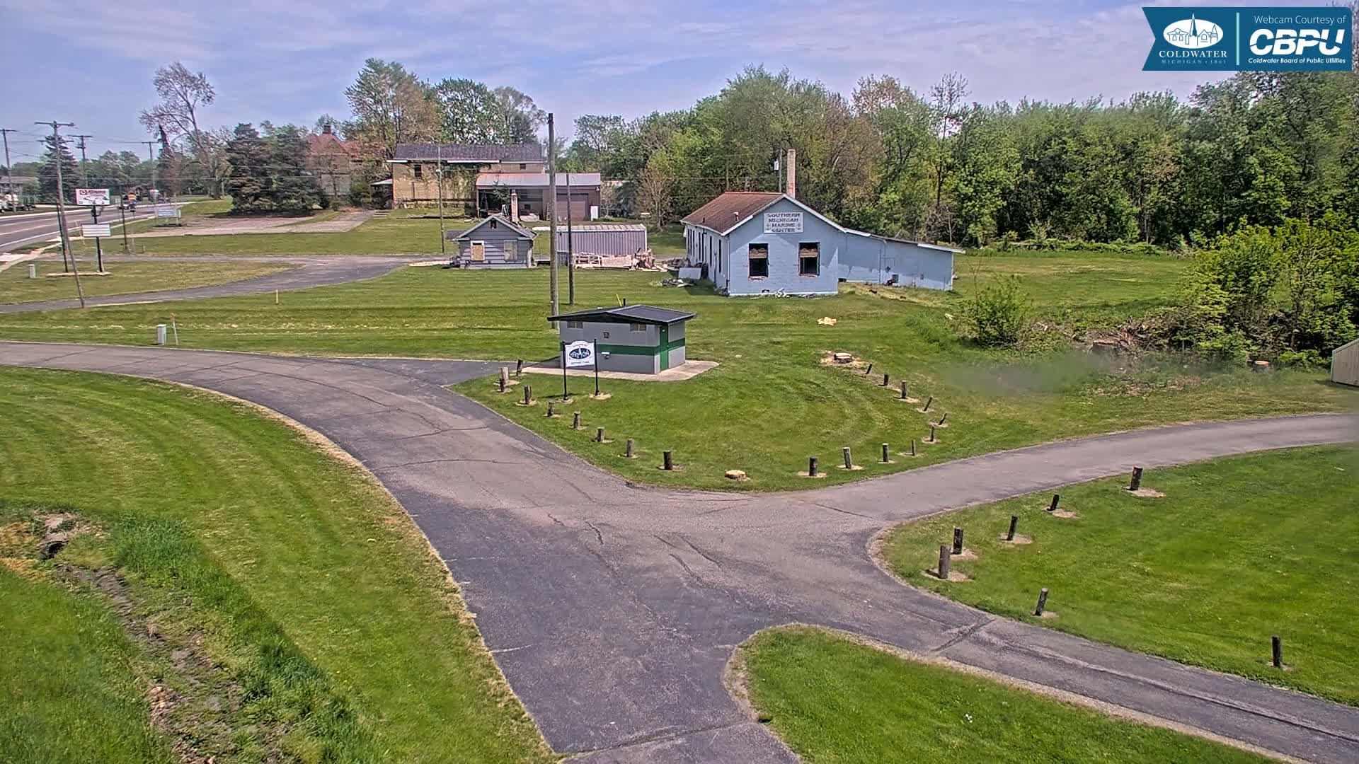 A paved pathway curves through a grassy area with several small buildings and trees under a partly sunny sky.