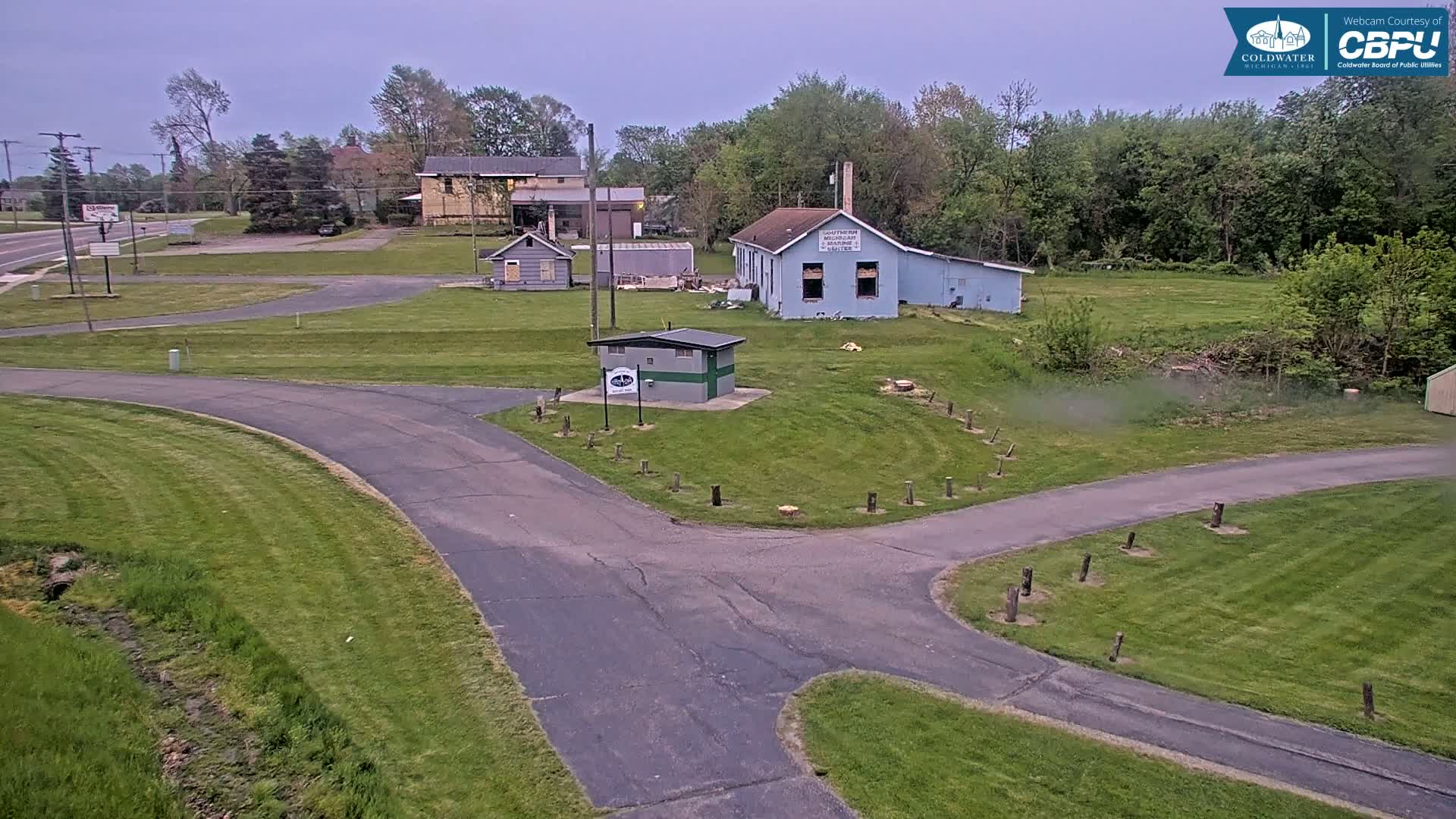 A paved roadway curves through a grassy area with several small buildings and trees under an overcast sky.