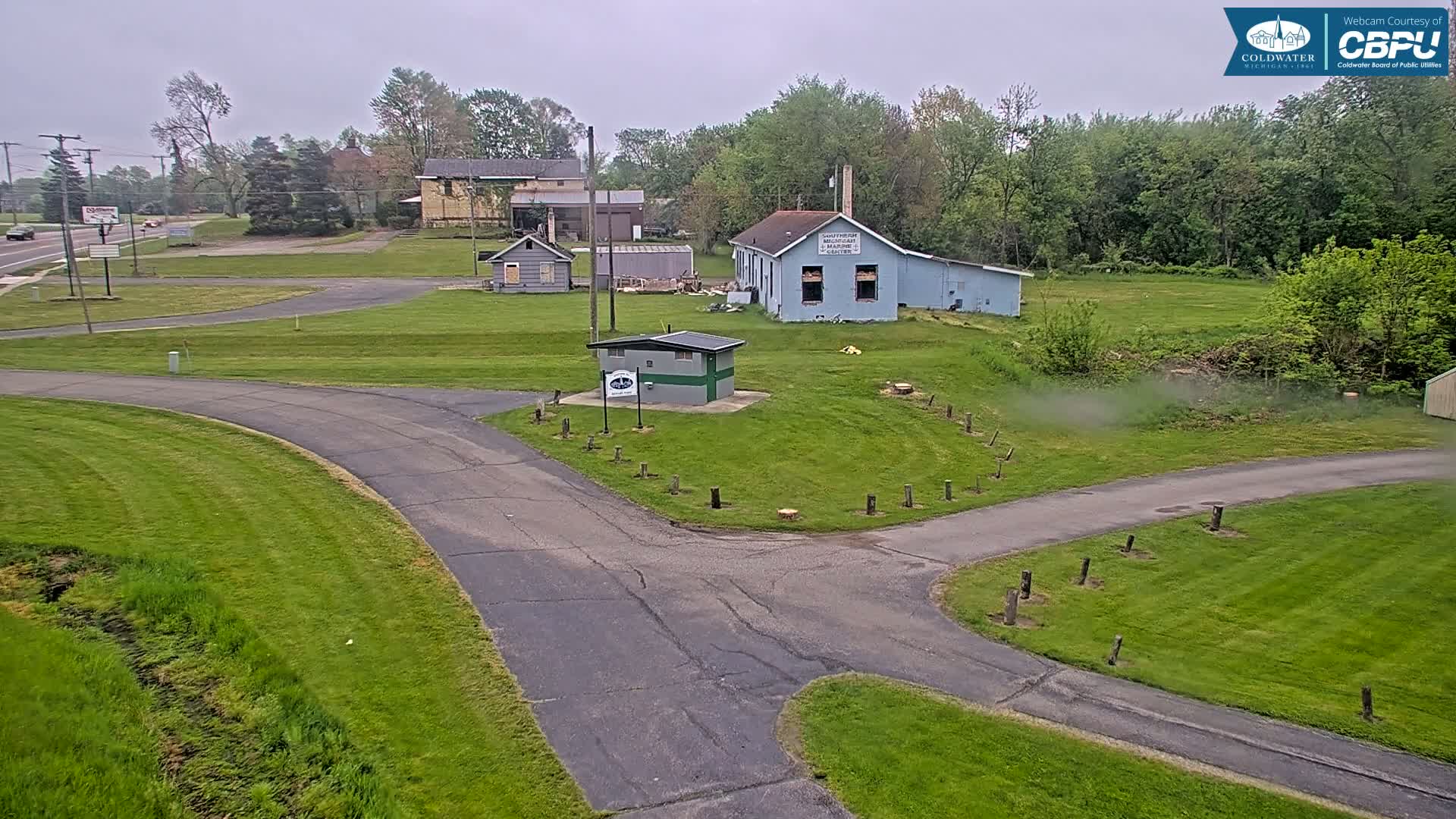 A paved roadway curves through a grassy area with several small buildings and trees under an overcast sky.