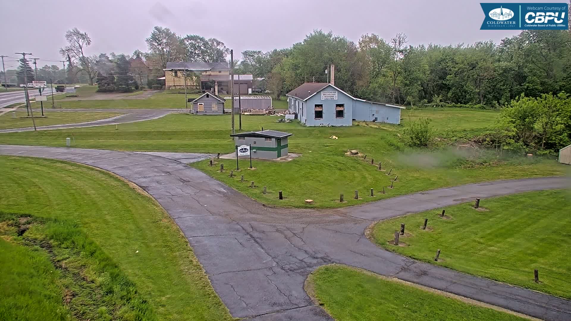 A paved roadway curves through a grassy area with several small buildings and trees under an overcast sky.