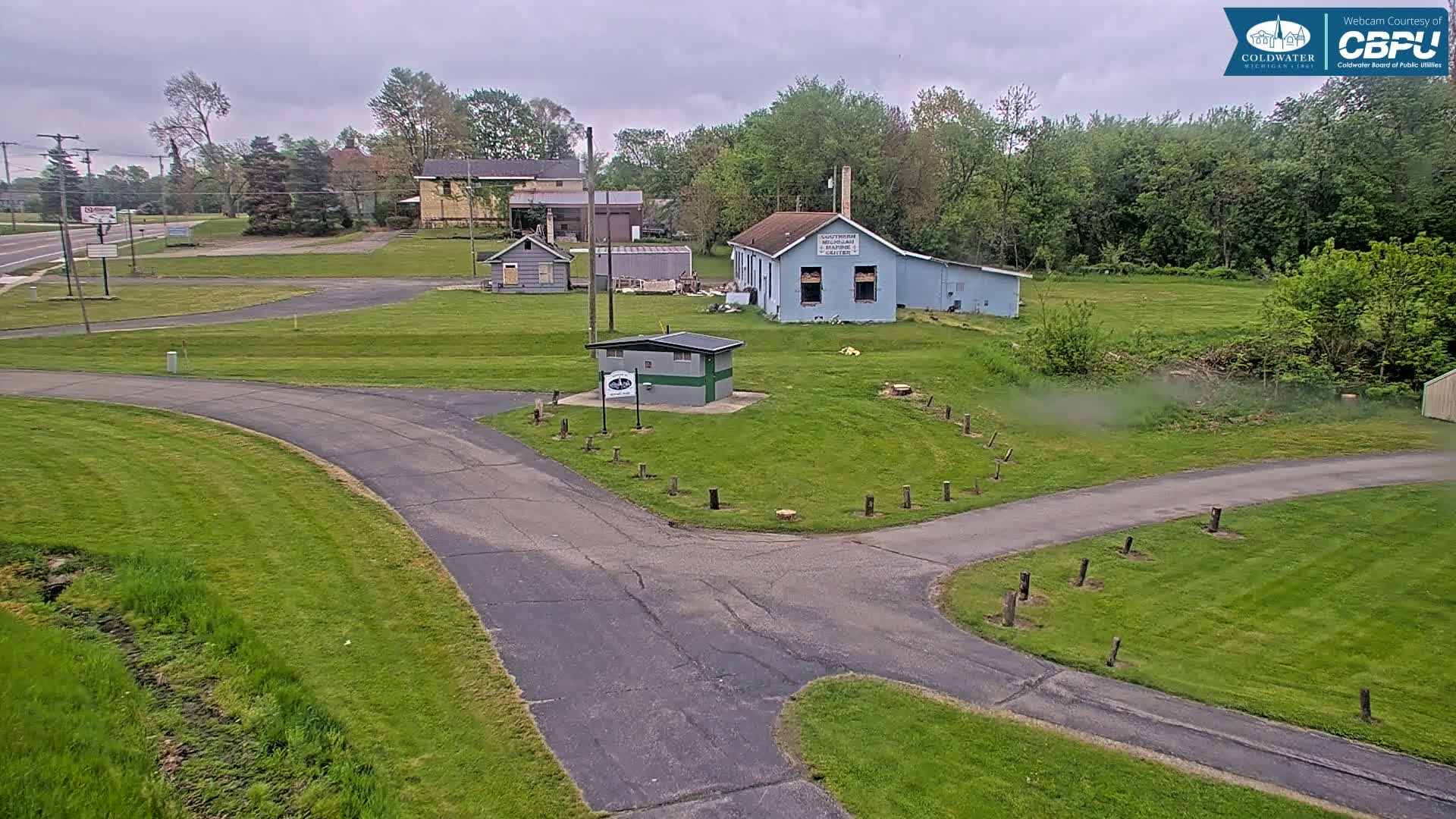A paved roadway intersects a grassy area with several small buildings and trees under an overcast sky.