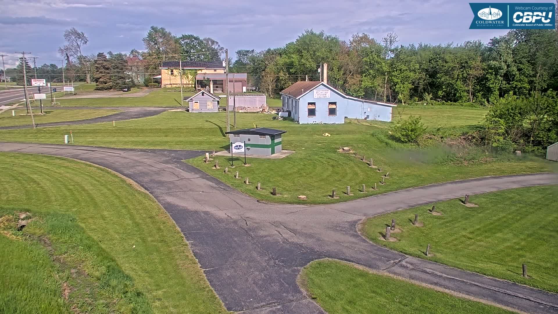 A paved roadway curves through a grassy area with several small buildings and a wooded backdrop under a partly cloudy sky.