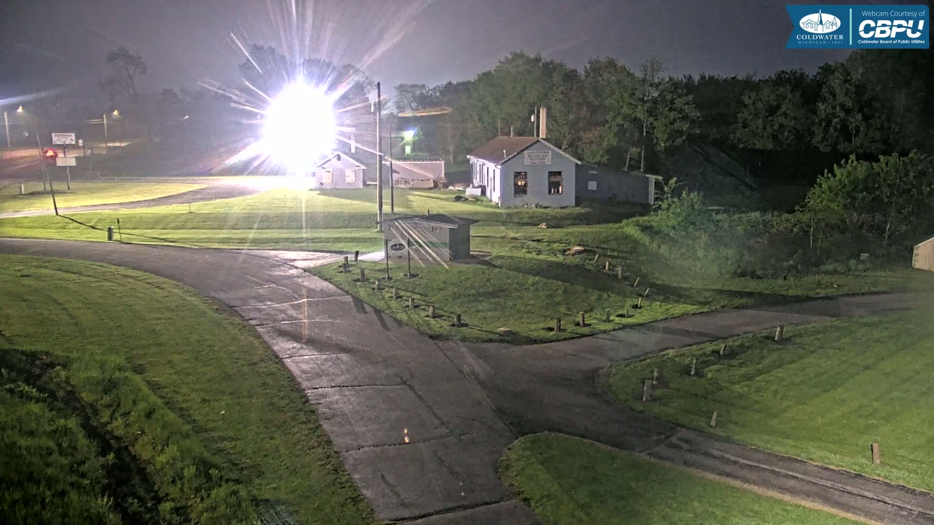 A nighttime view of a paved intersection surrounded by grassy areas and a few buildings, illuminated by bright lights under slightly hazy conditions.
