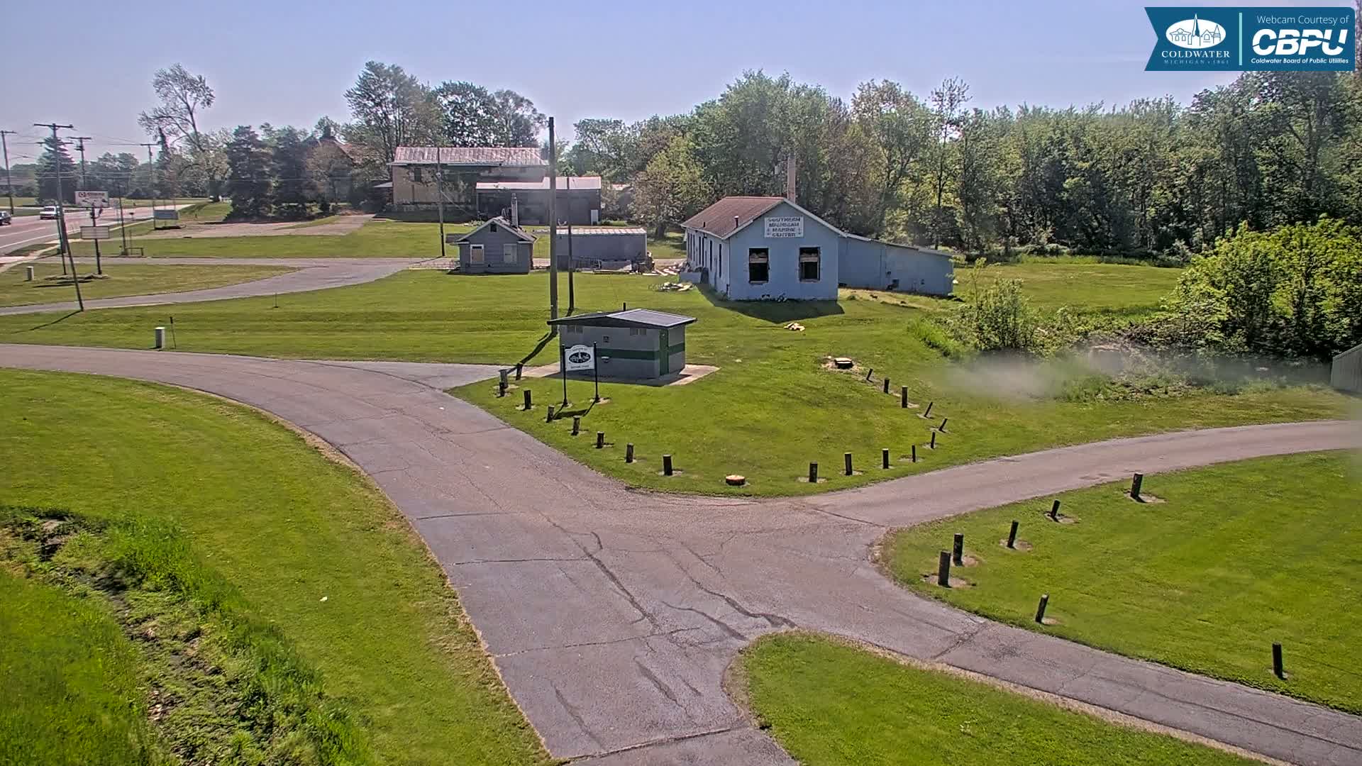 A paved road curves through a grassy area with several small buildings and trees under a clear, sunny sky.