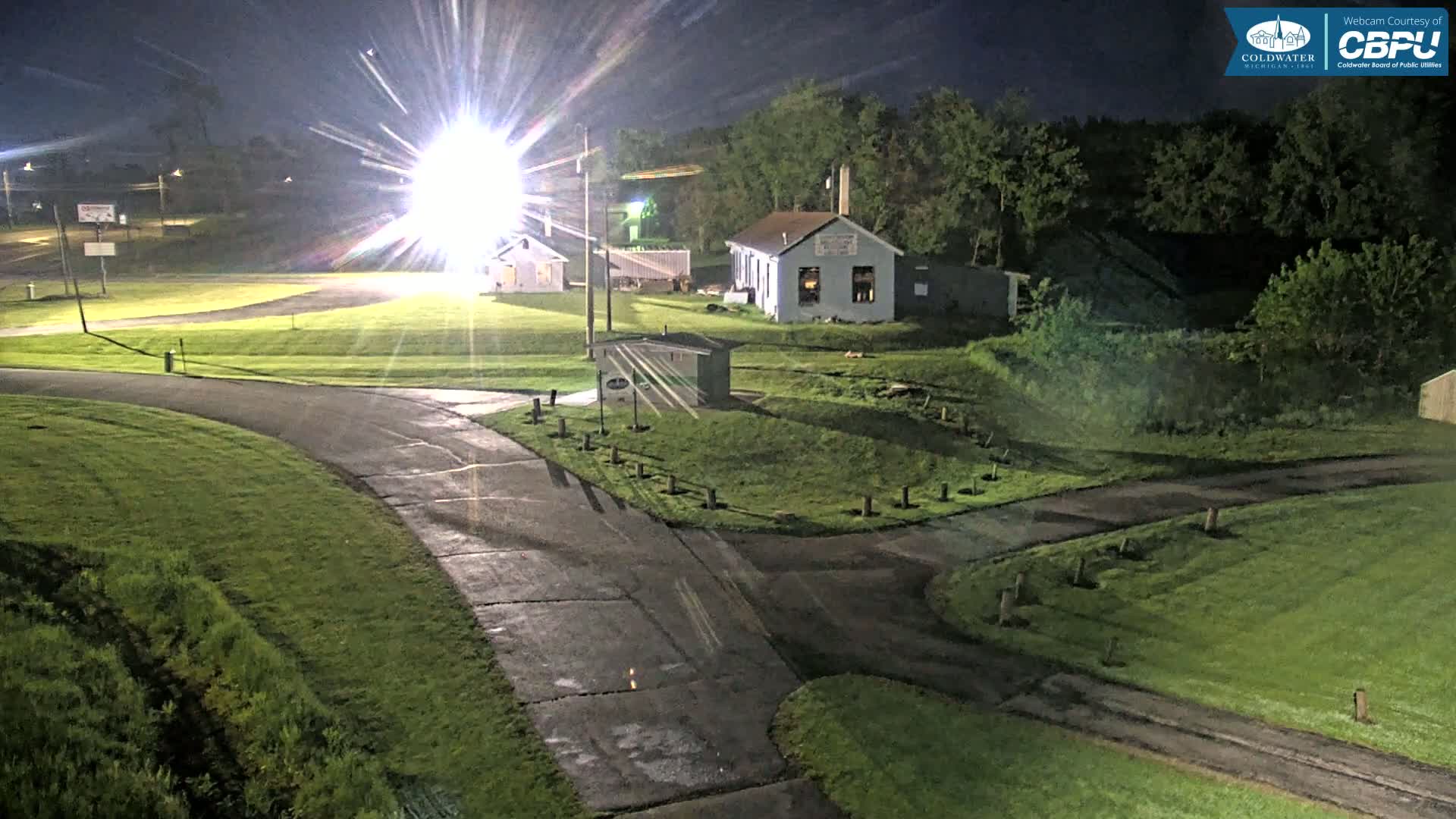 At night, under bright artificial light, a paved road curves through a grassy area with small buildings and trees in the background.