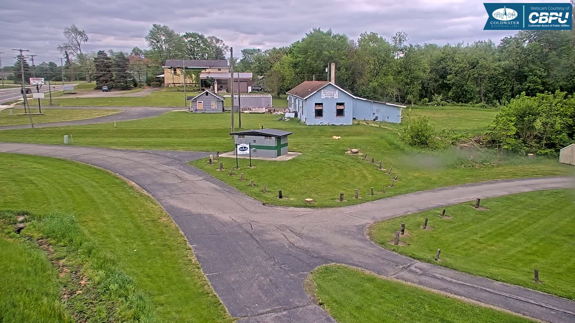A paved road intersects with a grassy area containing several small buildings under a cloudy sky.