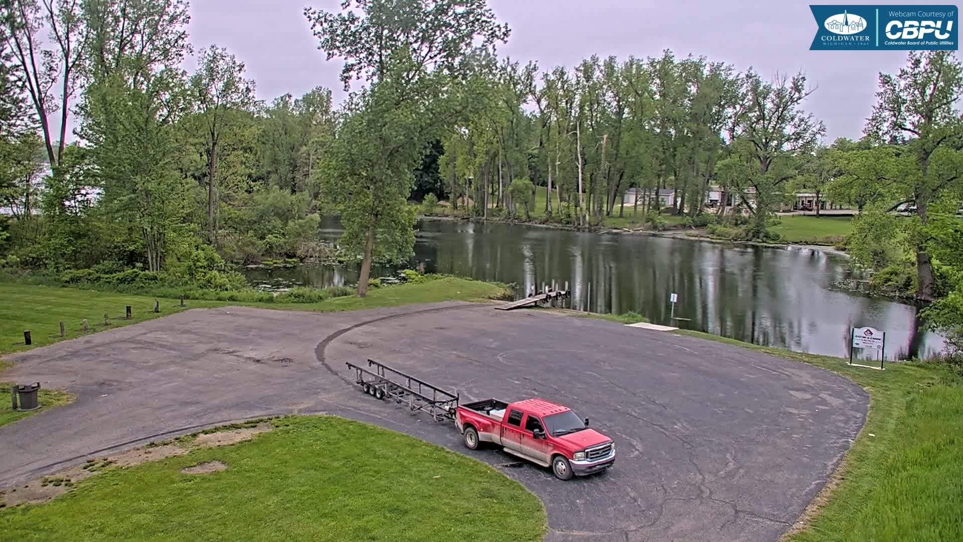 A red pickup truck with a boat trailer is parked in an empty boat launch area next to a calm body of water under an overcast sky, surrounded by lush green trees.