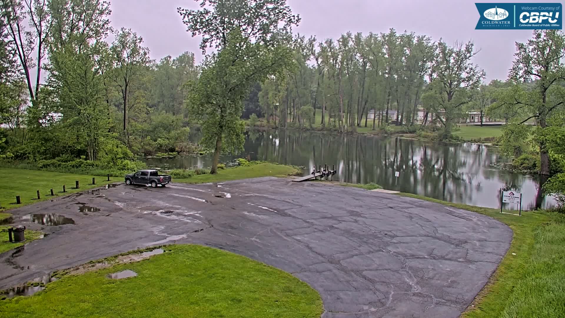 A dark-colored pickup truck is parked in a wet, cracked asphalt lot overlooking a calm lake surrounded by lush green trees under an overcast sky.