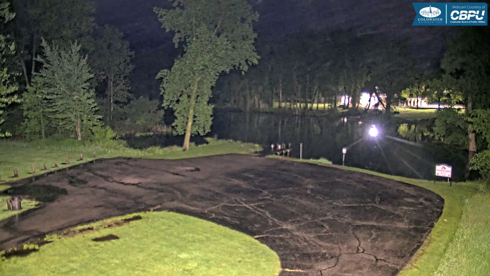 A dark asphalt parking lot next to a calm body of water, surrounded by trees at night.