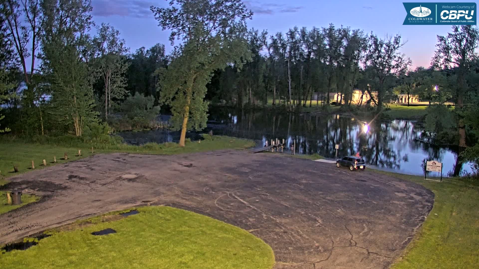 A dark-colored SUV is parked in a small asphalt lot next to a calm body of water, surrounded by trees under a twilight sky.