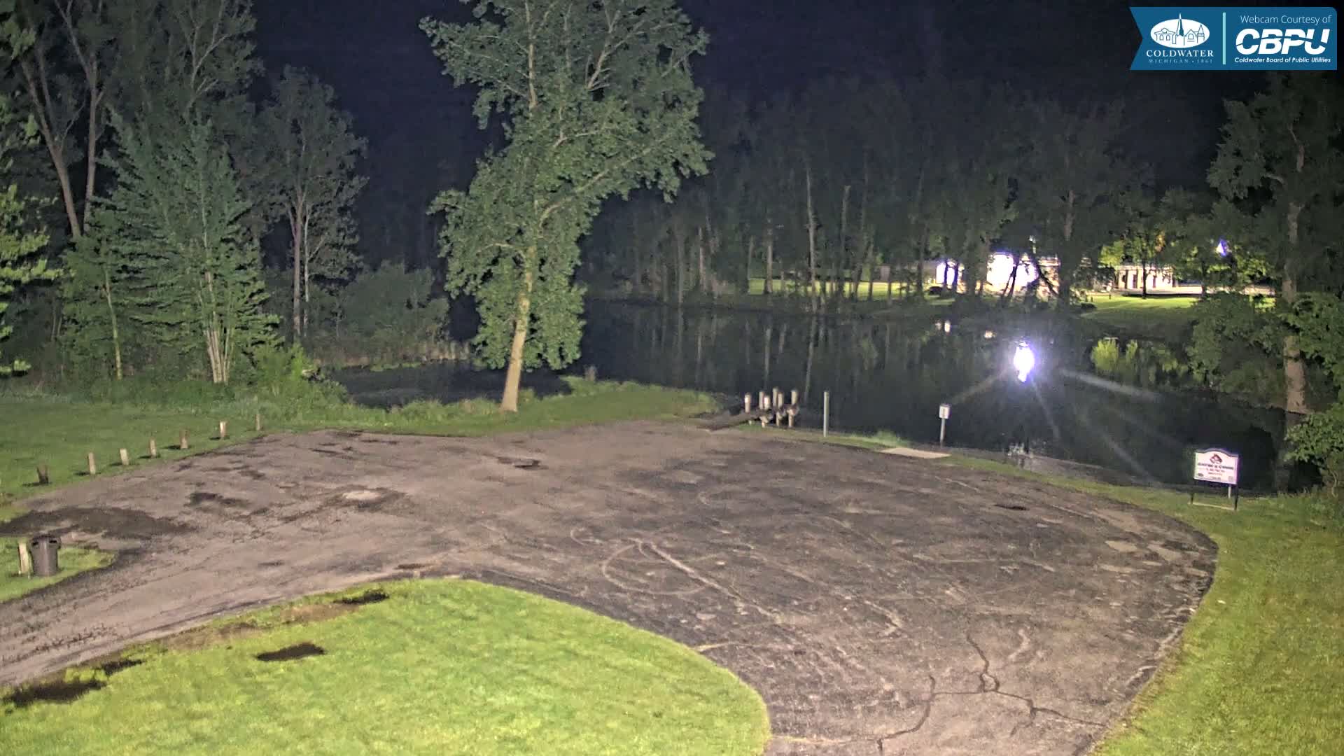 A nighttime view of a dark, empty asphalt boat launch area next to a calm body of water surrounded by trees.