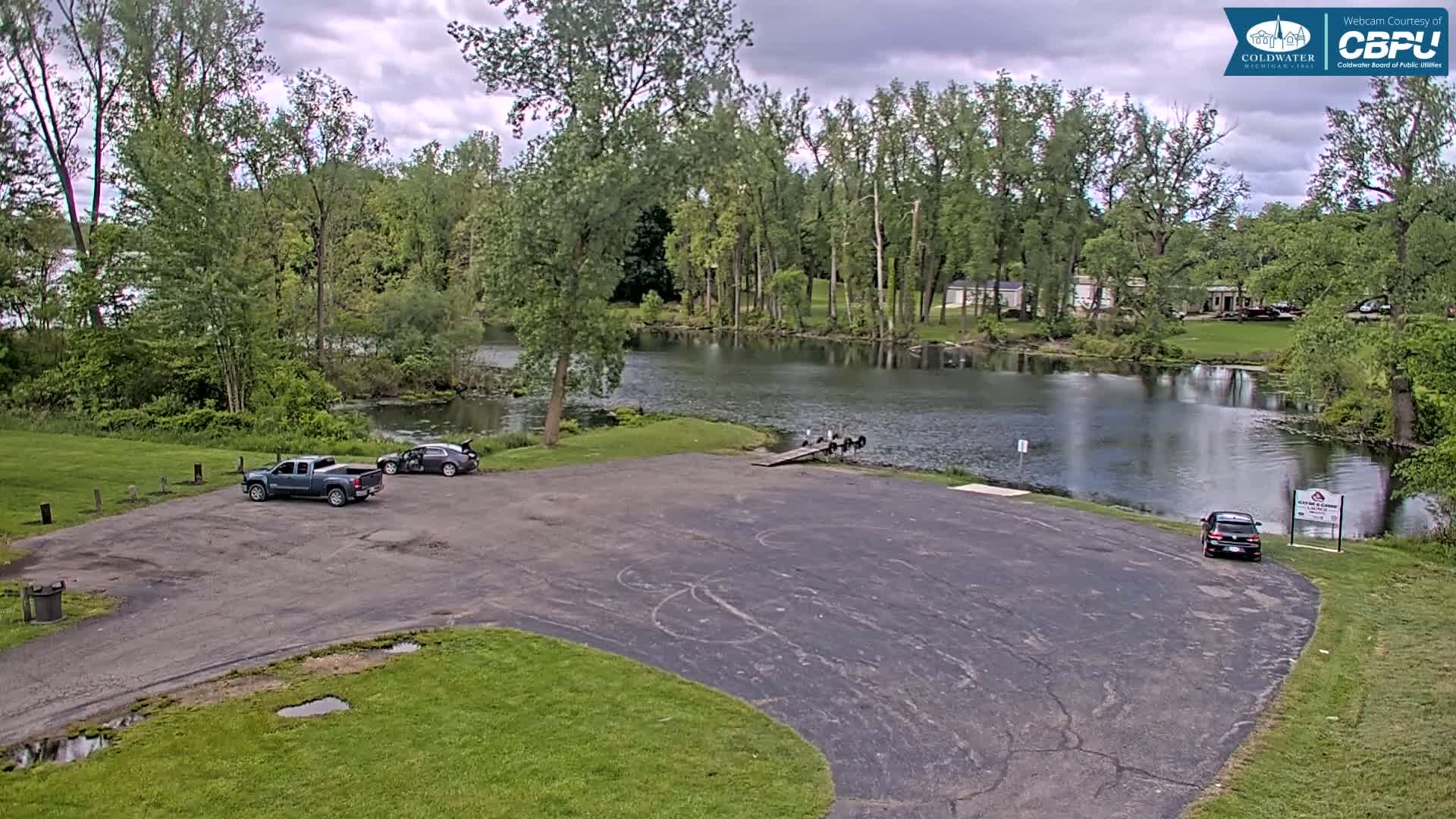 A mostly sunny day shows an empty parking lot next to a calm body of water, lined with lush green trees, with two vehicles parked and a boat ramp visible.