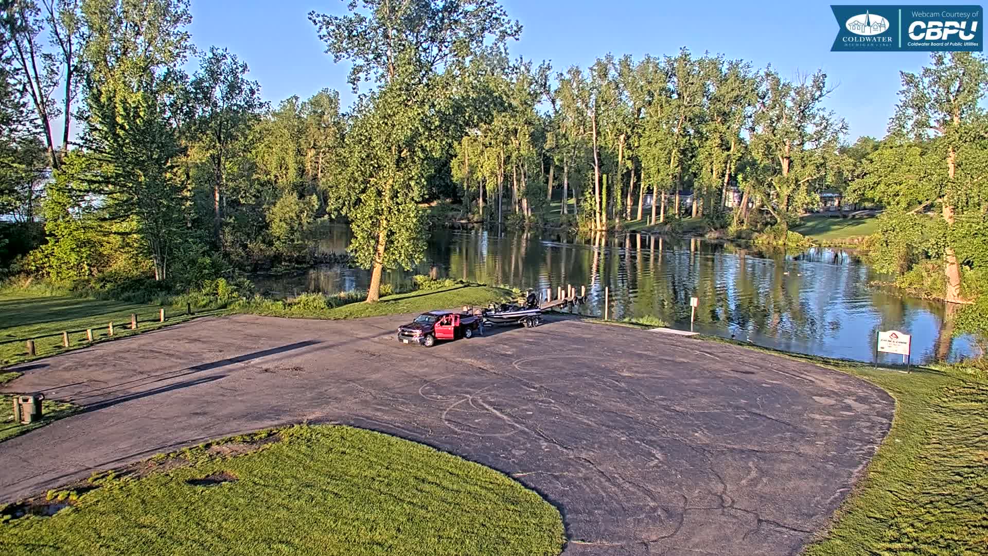 A red truck and fishing boat are parked in an empty asphalt lot next to a calm body of water lined with lush green trees under a clear, sunny sky.