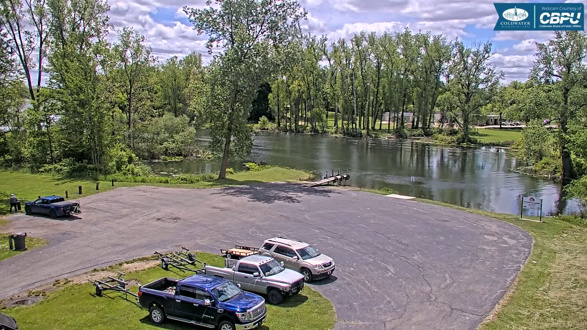 A paved parking lot with several trucks and boat trailers sits beside a calm river under a partly cloudy sky, surrounded by lush green trees.