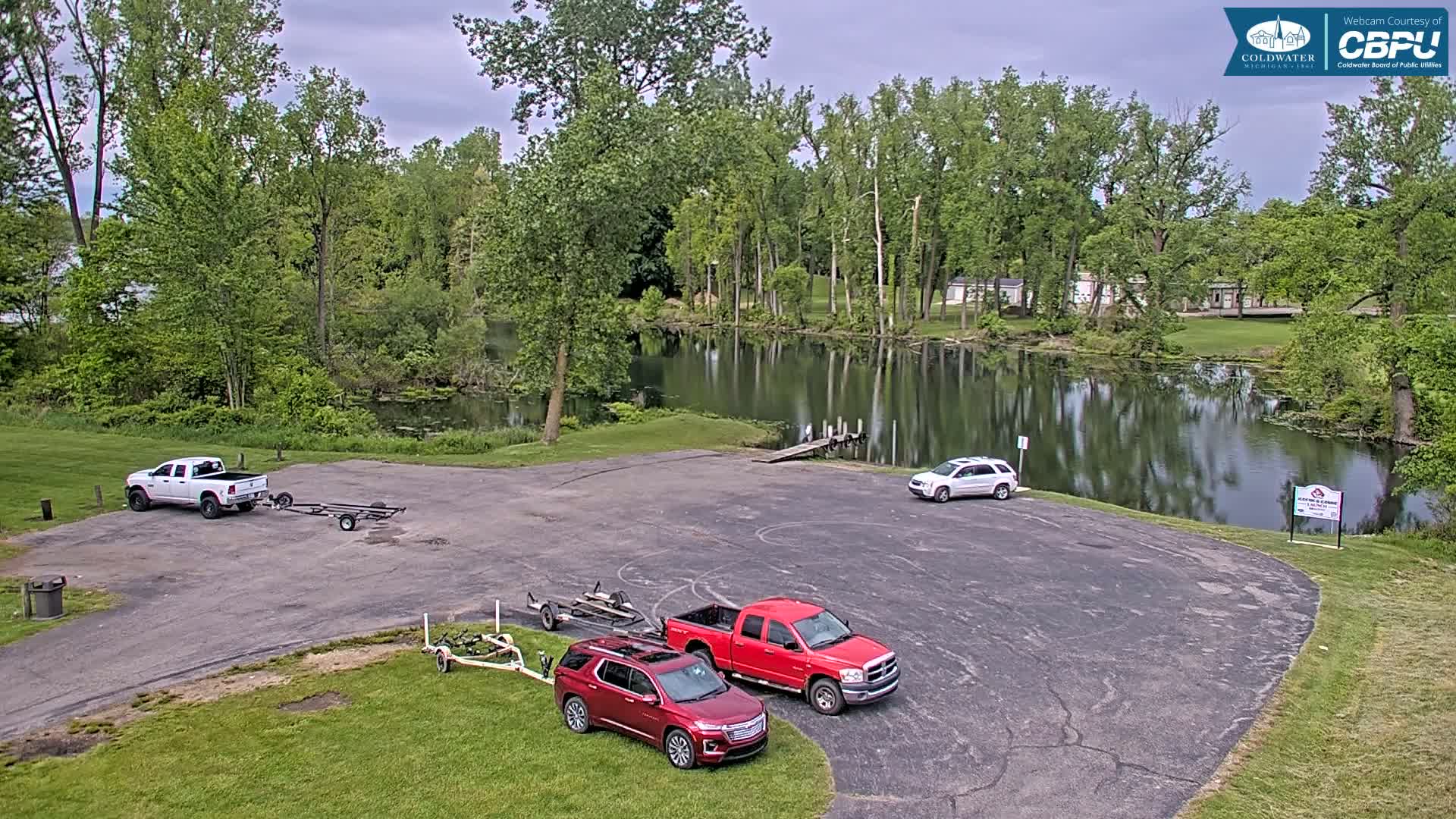 A paved boat launch area with several trucks and SUVs, some towing boat trailers, overlooks a calm pond surrounded by lush green trees under a partly cloudy sky.