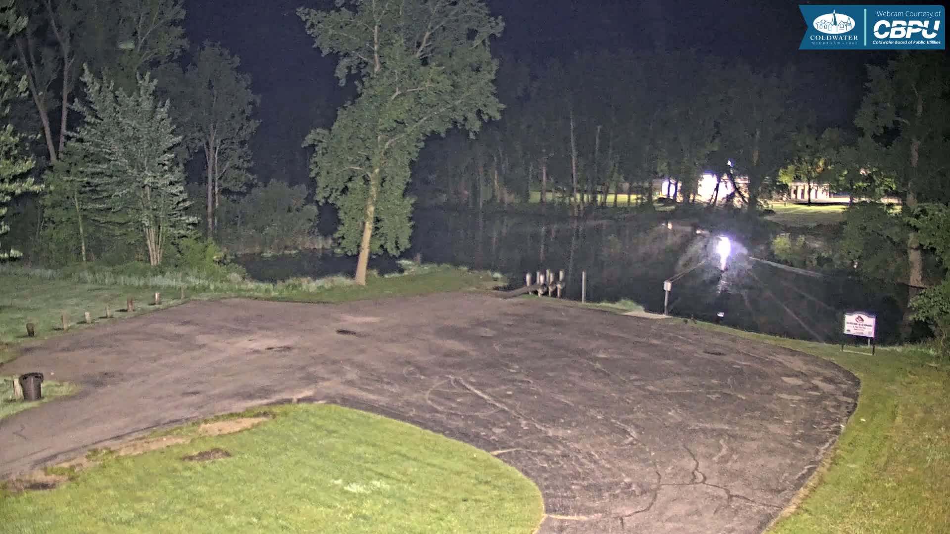 An empty asphalt parking lot at night next to a calm body of water surrounded by trees under nighttime lighting.