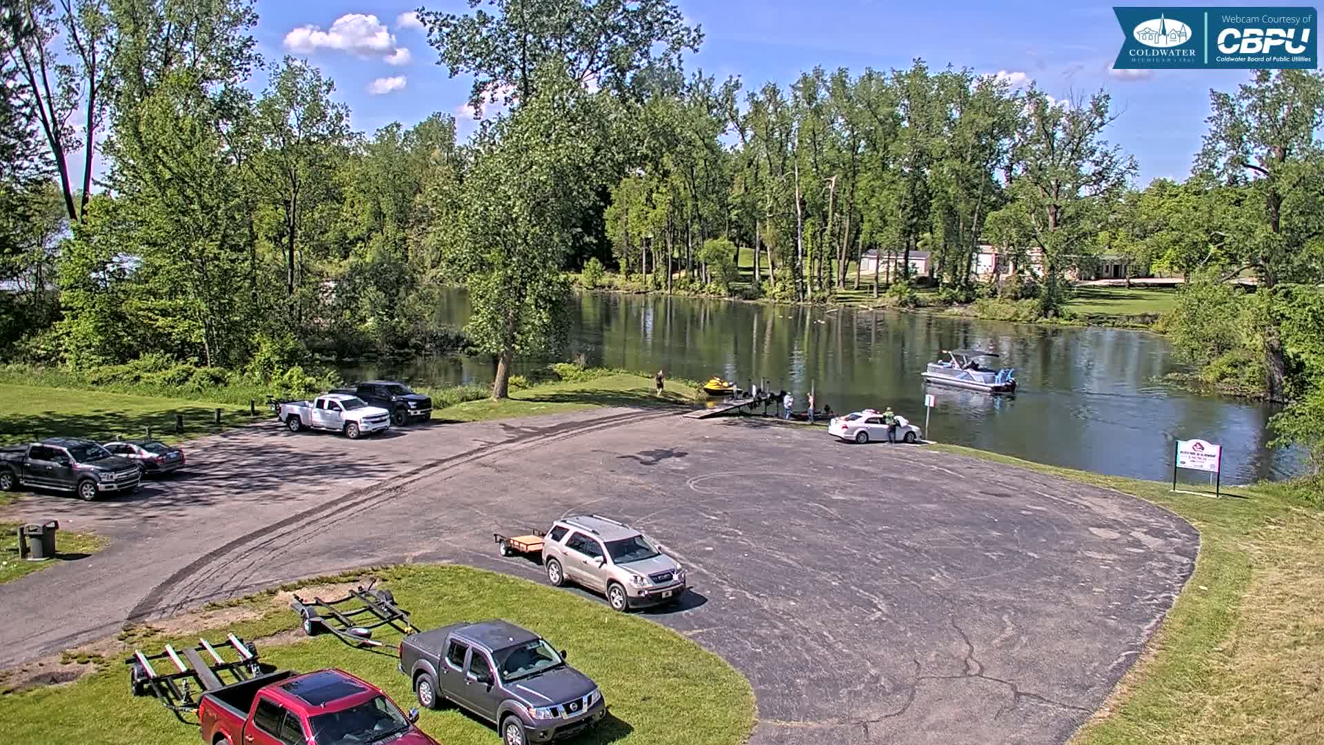 A sunny day at a boat launch with several vehicles parked in the lot, a boat on the water, and people near a dock.