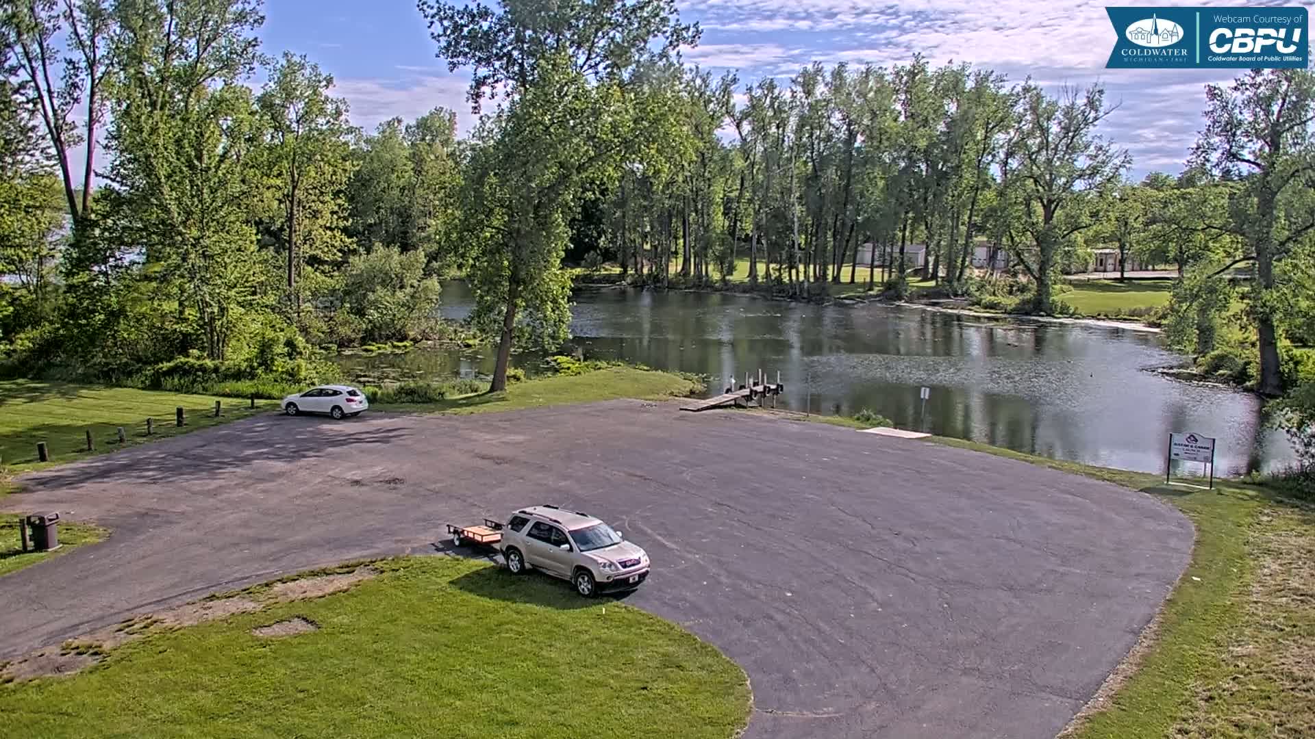 An empty parking lot with two cars and a boat trailer overlooks a calm river lined with lush green trees under a partly sunny sky.