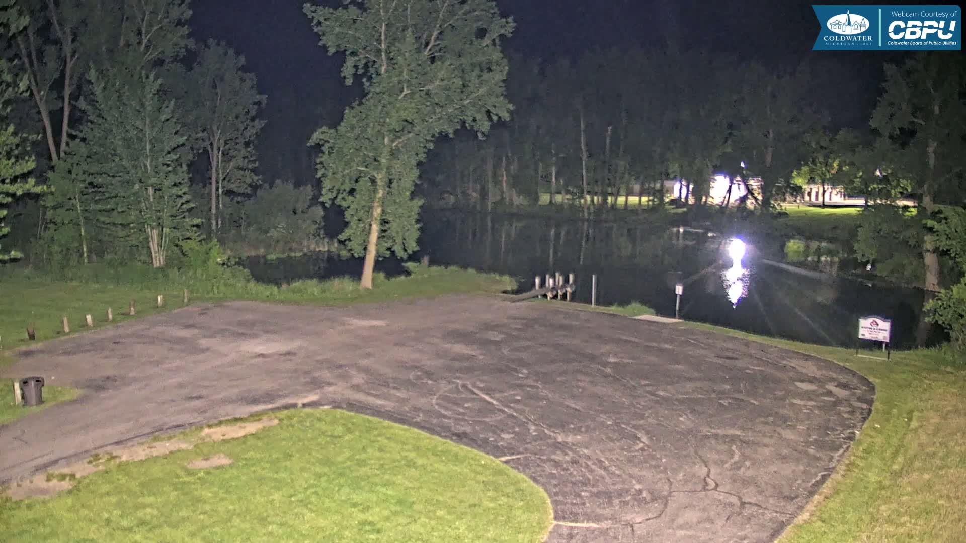A nighttime view of an empty asphalt parking lot next to a calm body of water surrounded by trees under a dark sky.