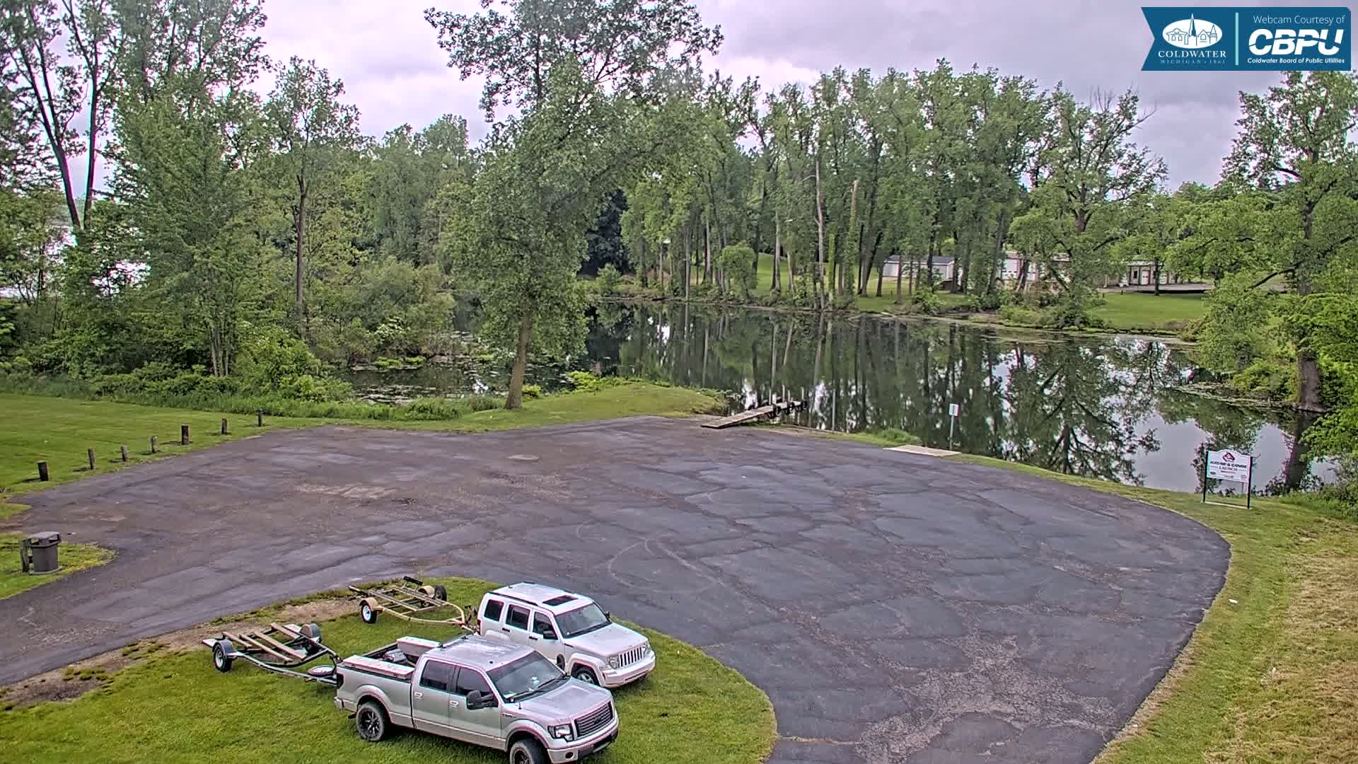 A paved boat launch area with two trucks and trailers sits beside a calm body of water surrounded by lush green trees under an overcast sky.