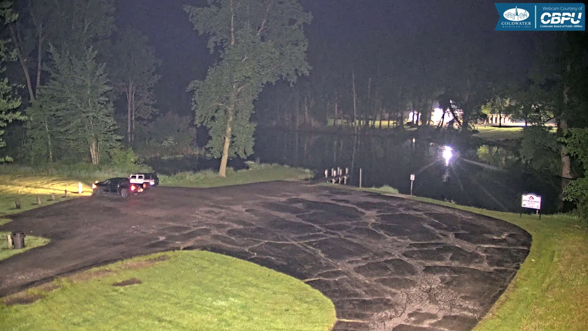 Two vehicles are parked in a cracked asphalt lot at night next to a calm body of water surrounded by trees.