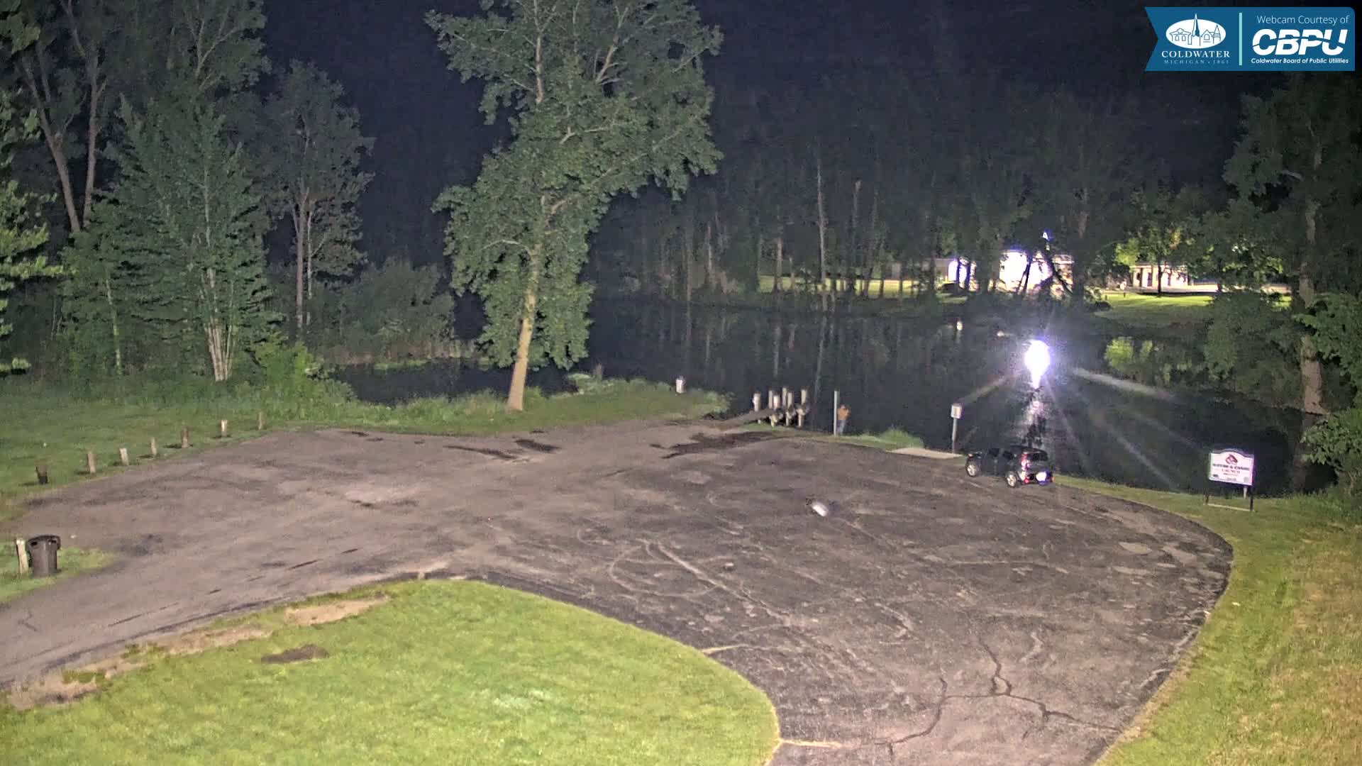 At night, a dark asphalt parking lot curves toward a calm body of water bordered by trees under a dark sky.