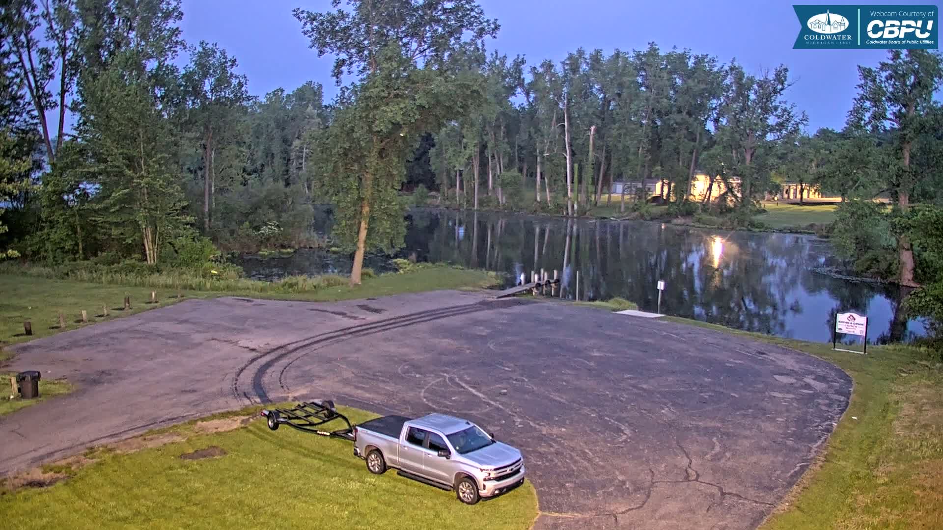 A silver pickup truck with a boat trailer is parked in a paved lot next to a calm, dark body of water surrounded by trees under a twilight sky.