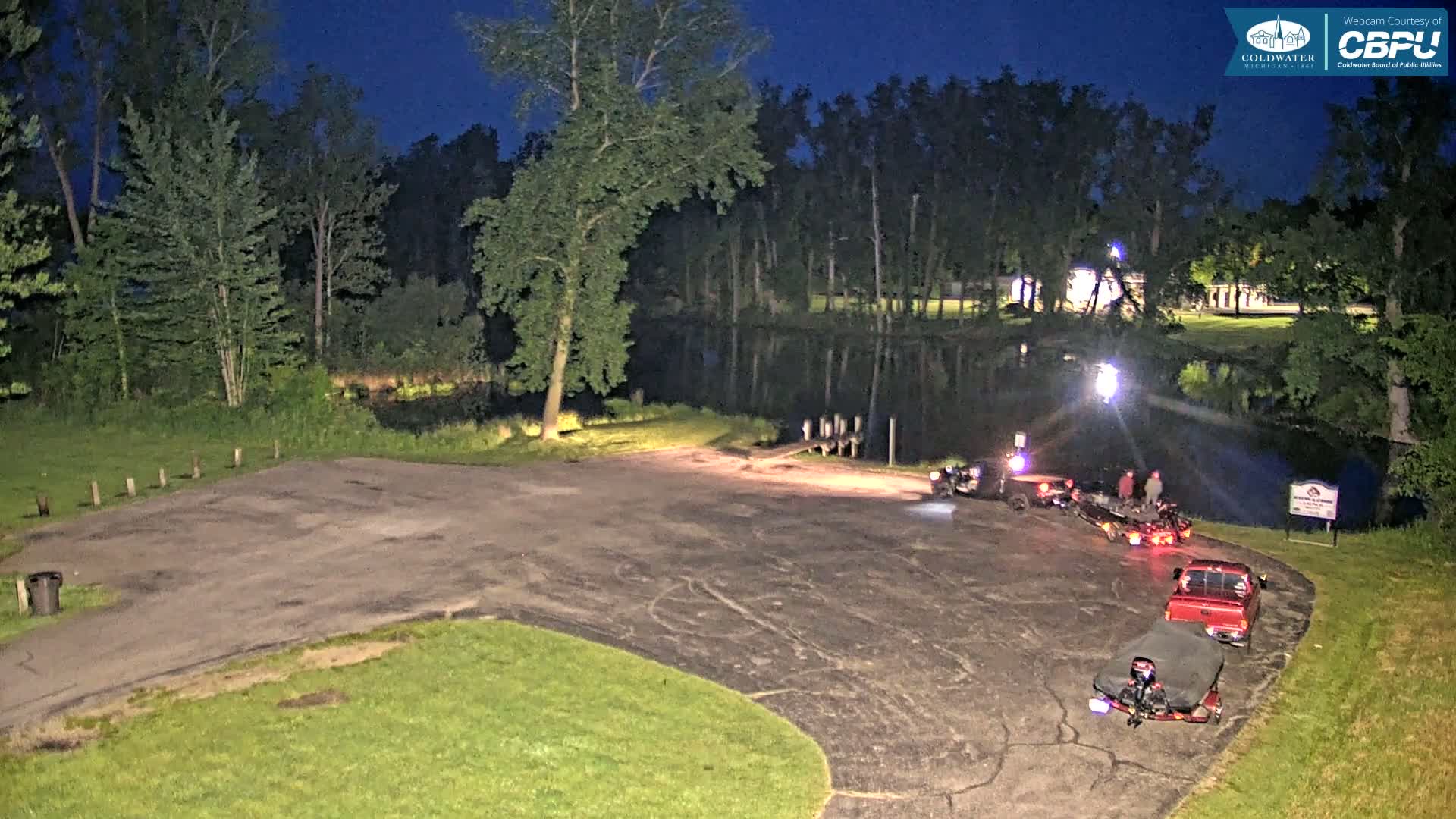 At night, several vehicles and people are gathered in a parking lot next to a calm body of water bordered by trees.
