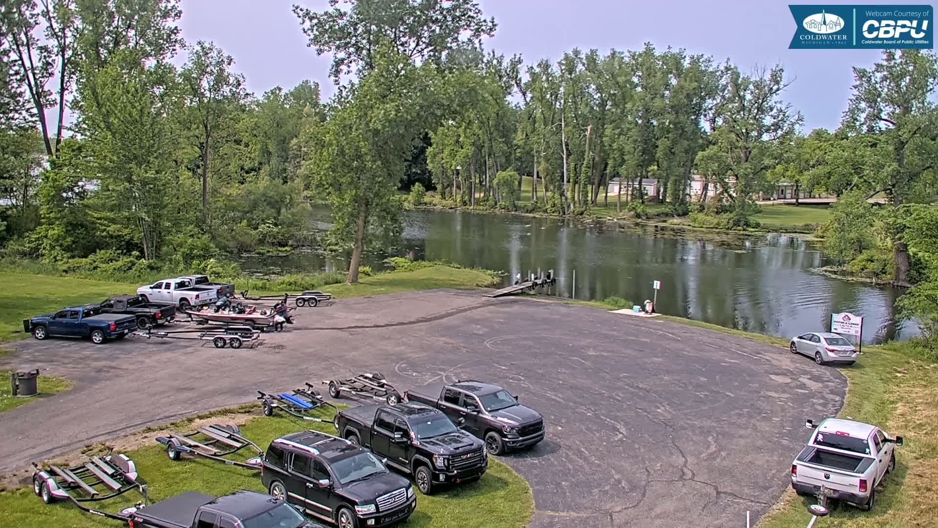 Several trucks and boats are parked in a paved lot next to a calm lake surrounded by lush green trees under a partly cloudy sky.
