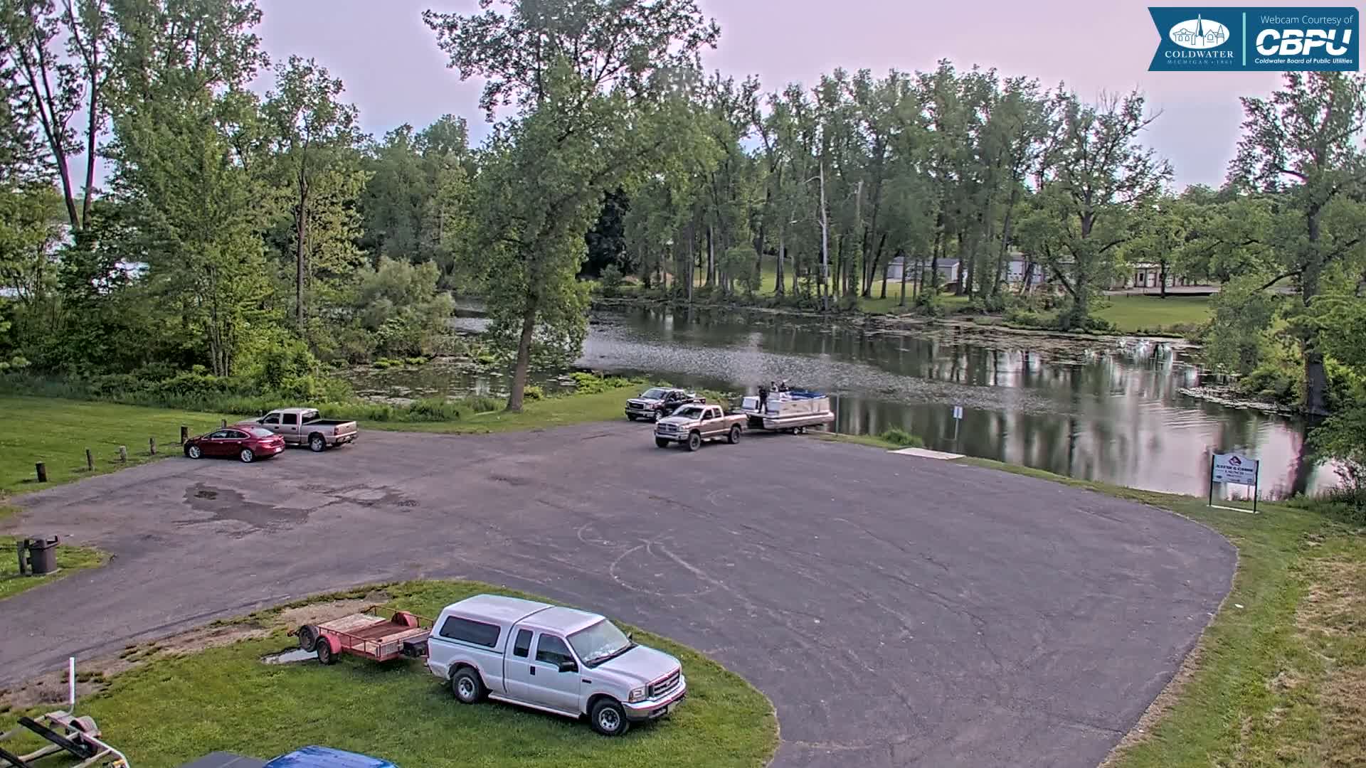 A paved parking lot with several trucks and a pontoon boat near a calm lake surrounded by lush green trees under an overcast sky.