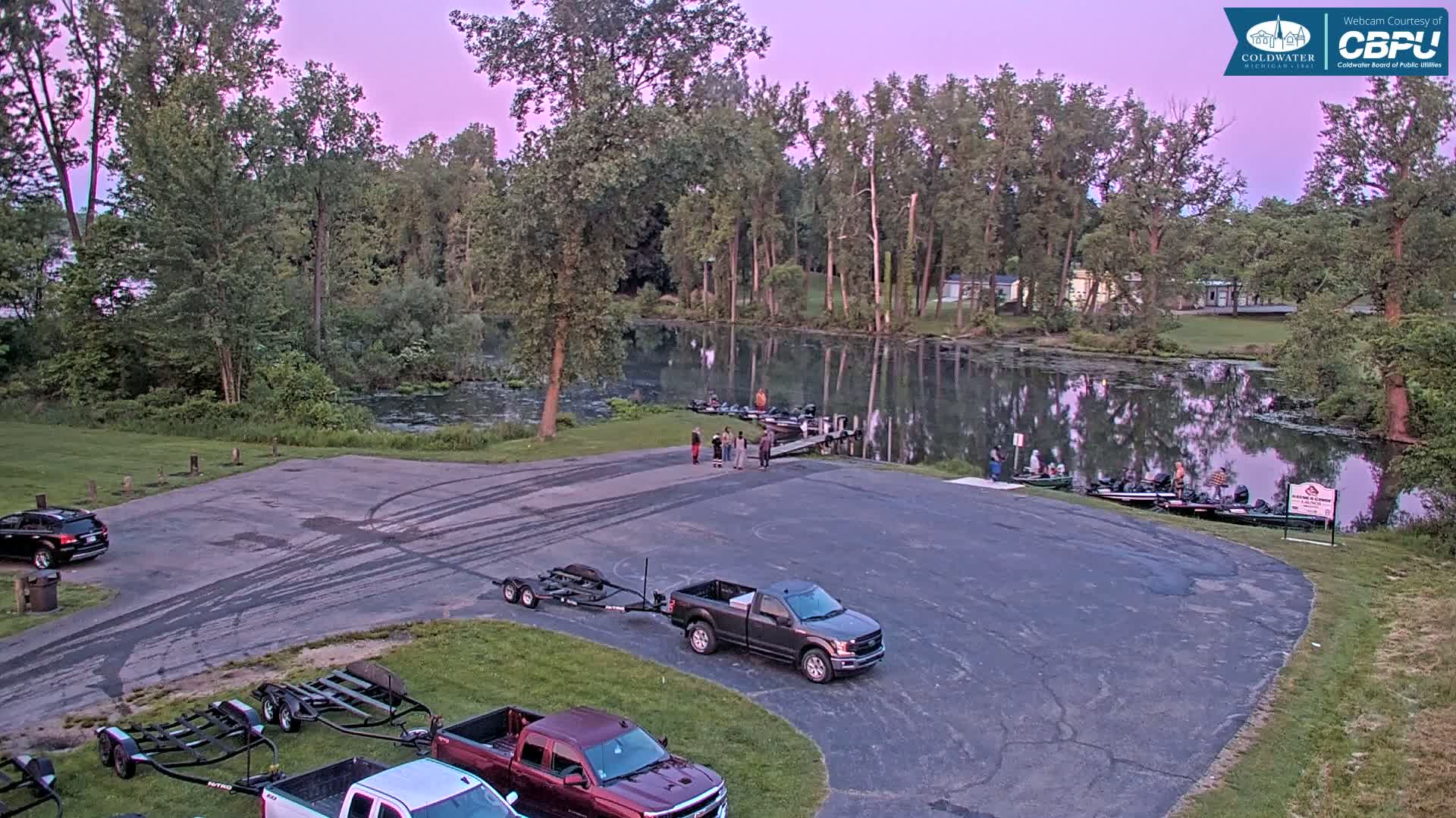 A calm, twilight-lit lake is seen from a high angle, showing a boat launch with several parked trucks and boat trailers, along with a few people near the water's edge.