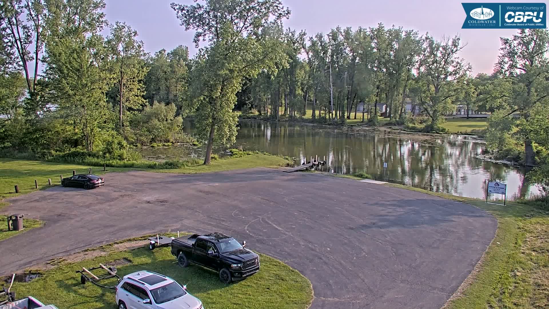 An empty parking lot next to a calm lake surrounded by lush green trees under a partly sunny sky.