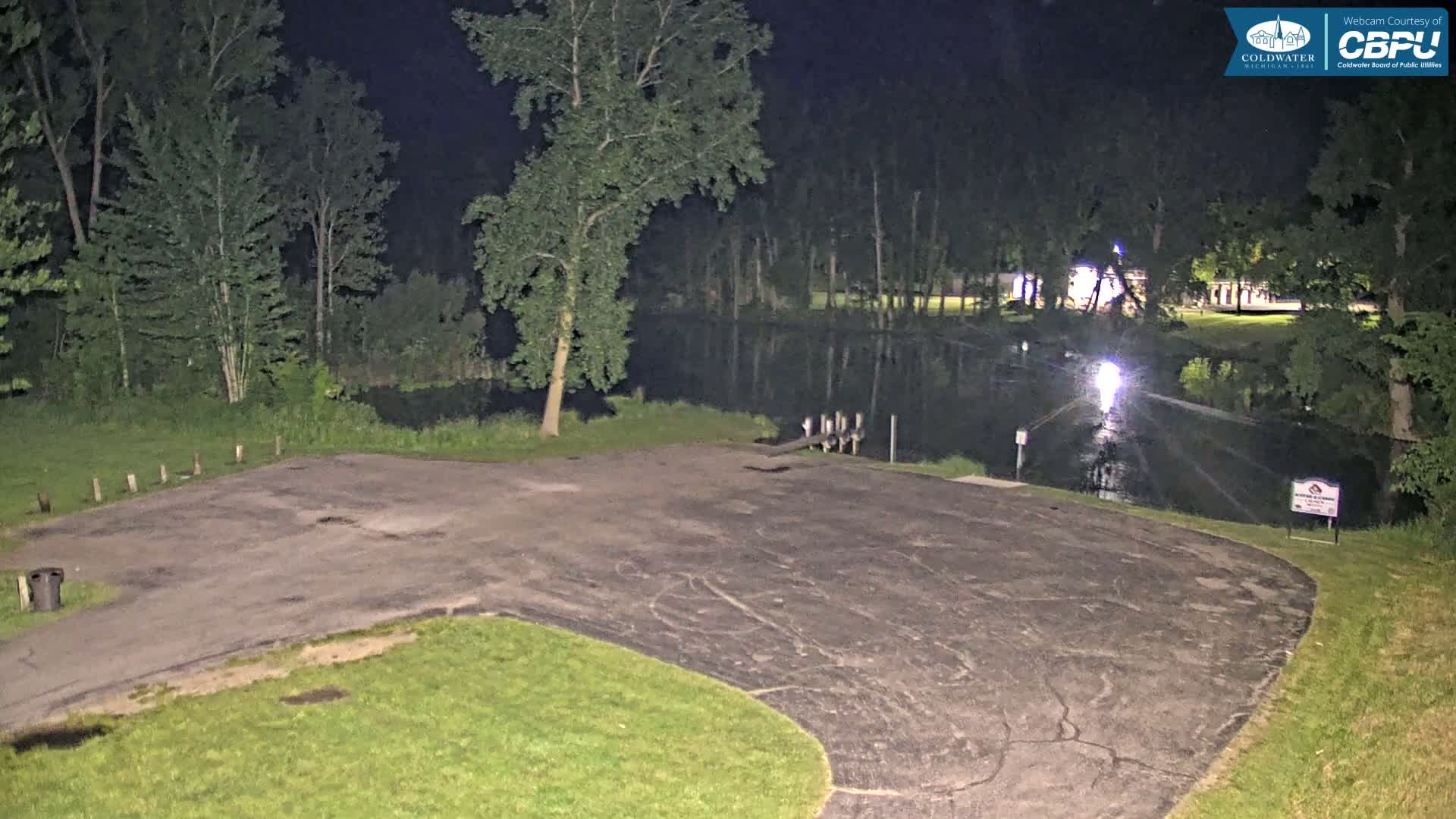 An empty, dark asphalt parking lot at night next to a calm body of water bordered by trees under nighttime lighting.