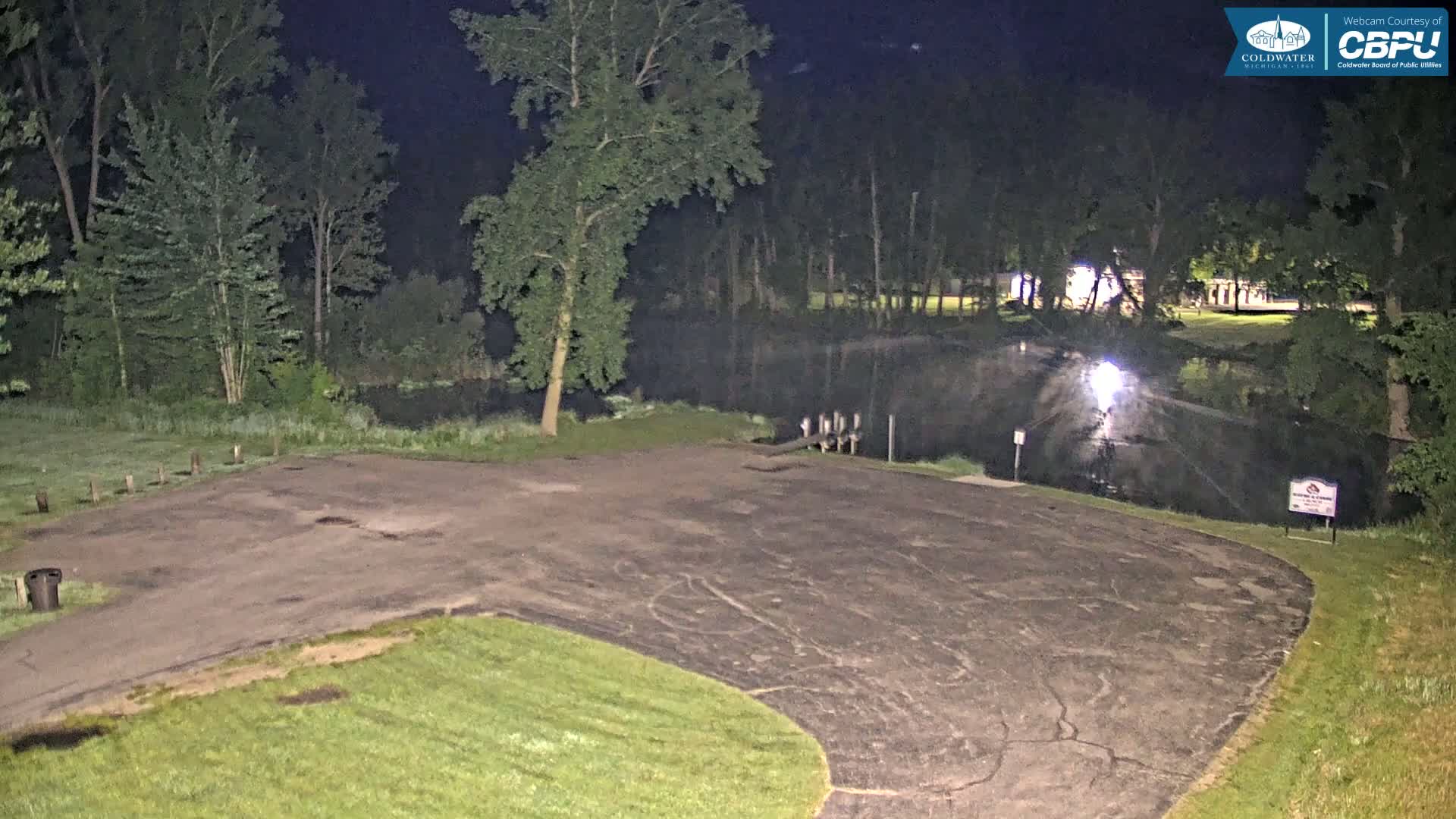 A nighttime view of an empty paved parking lot next to a calm body of water, surrounded by trees under dark, clear skies.