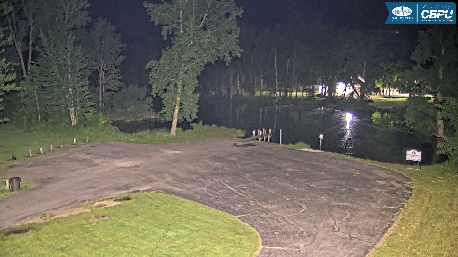 A nighttime view of an empty asphalt parking lot next to a calm body of water bordered by trees.