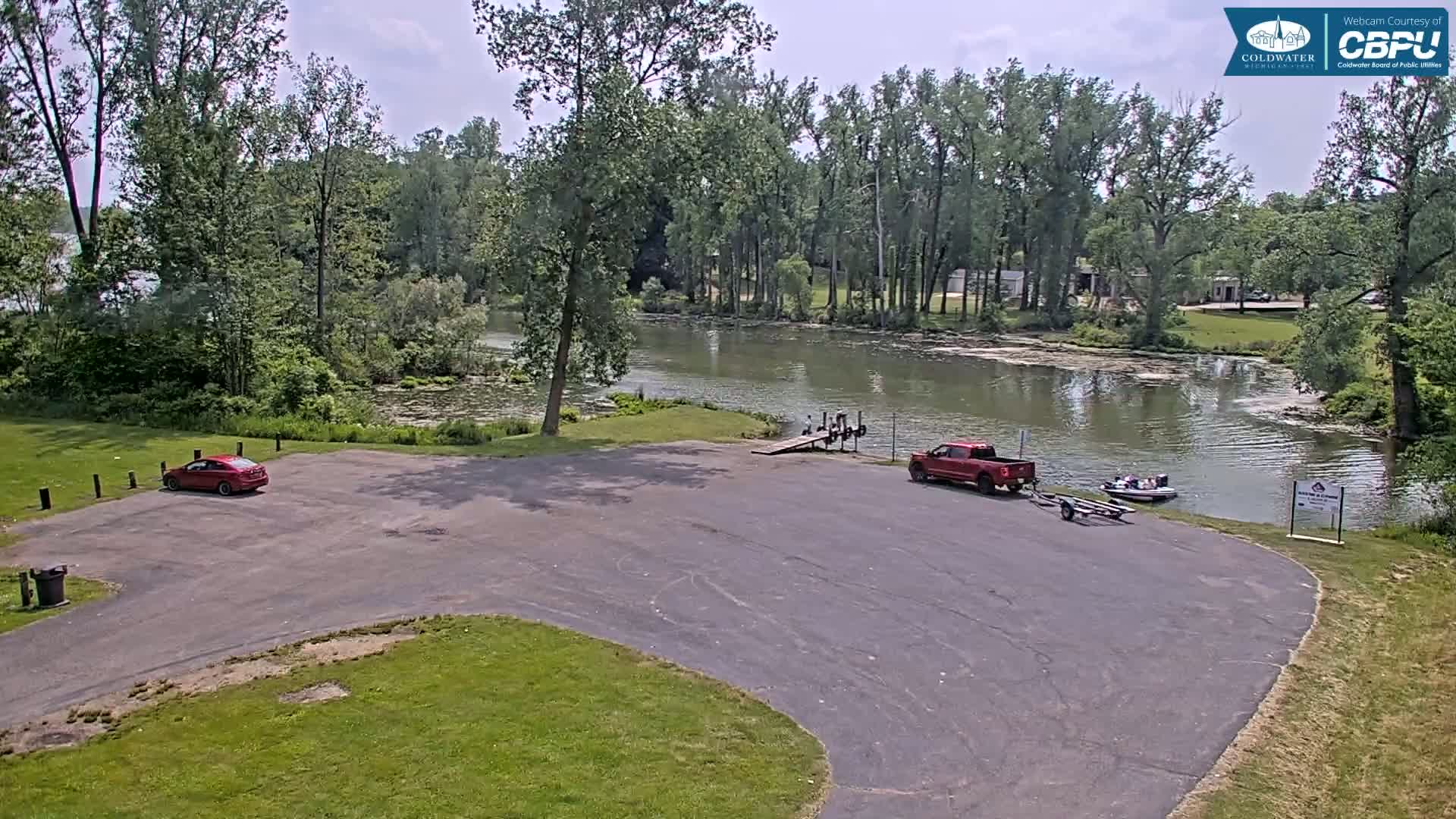 An empty parking lot next to a calm body of water, surrounded by lush green trees under a partly cloudy sky, with a red car and a red truck parked nearby, and a small boat on the water.