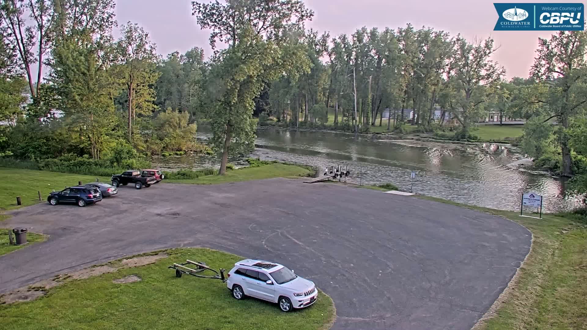 A parking lot with several vehicles is situated next to a calm river lined with lush green trees under a partly cloudy sky.