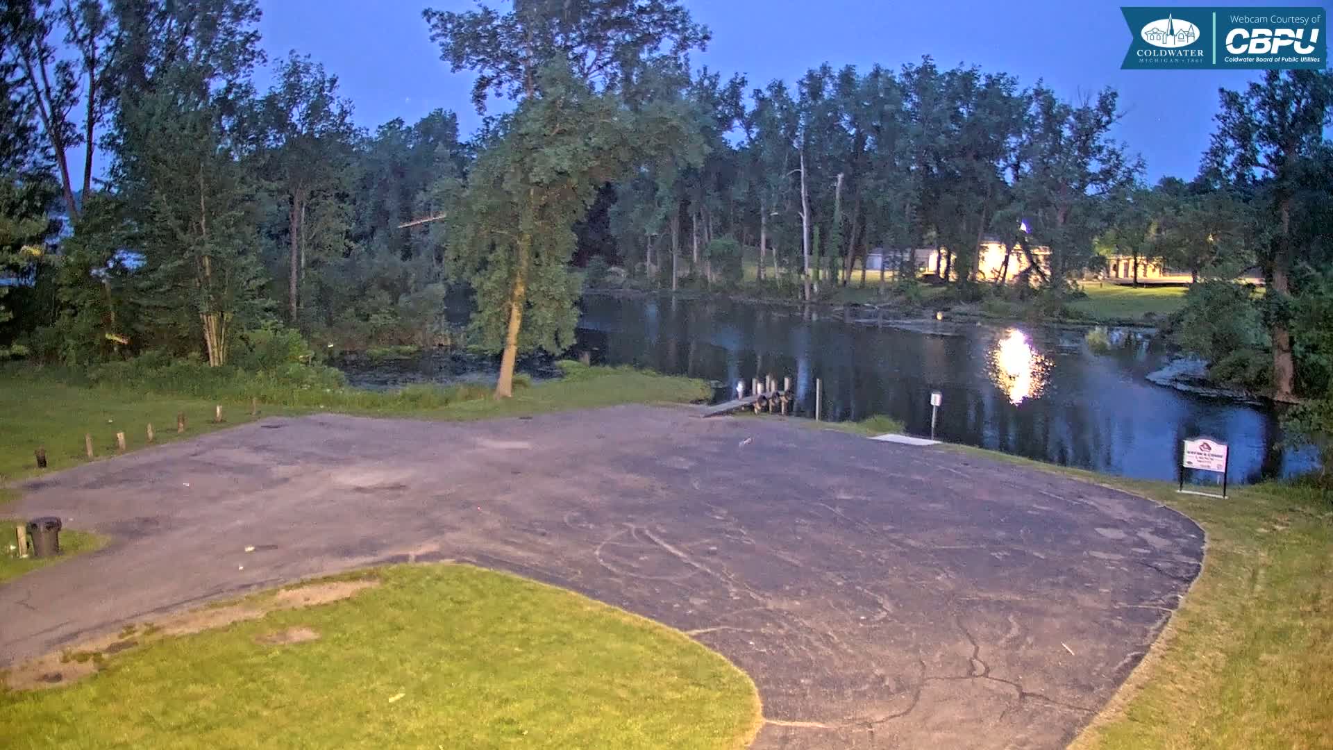 An empty asphalt parking lot next to a calm body of water under a twilight sky, bordered by lush green trees.