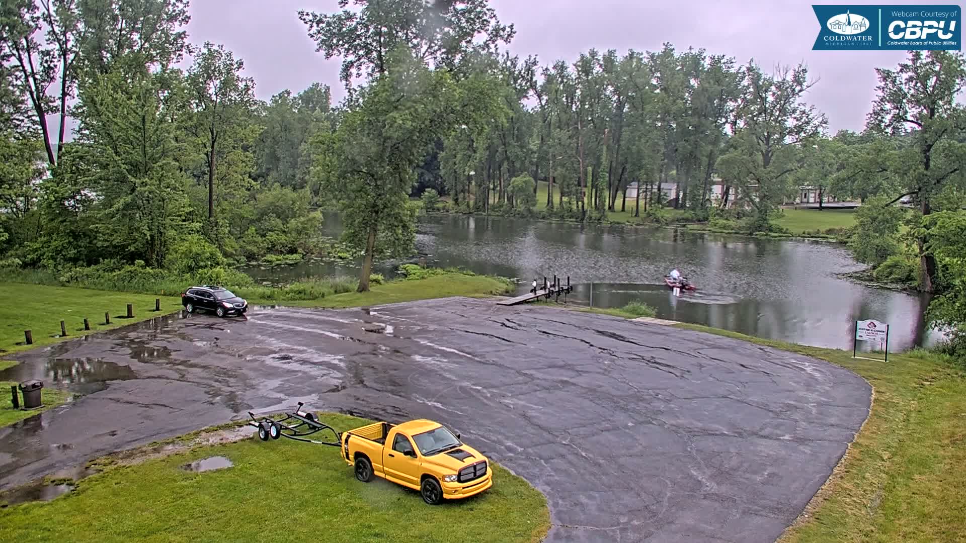 A yellow pickup truck and a dark-colored SUV are parked in a wet parking lot next to a calm lake surrounded by lush green trees under an overcast sky.