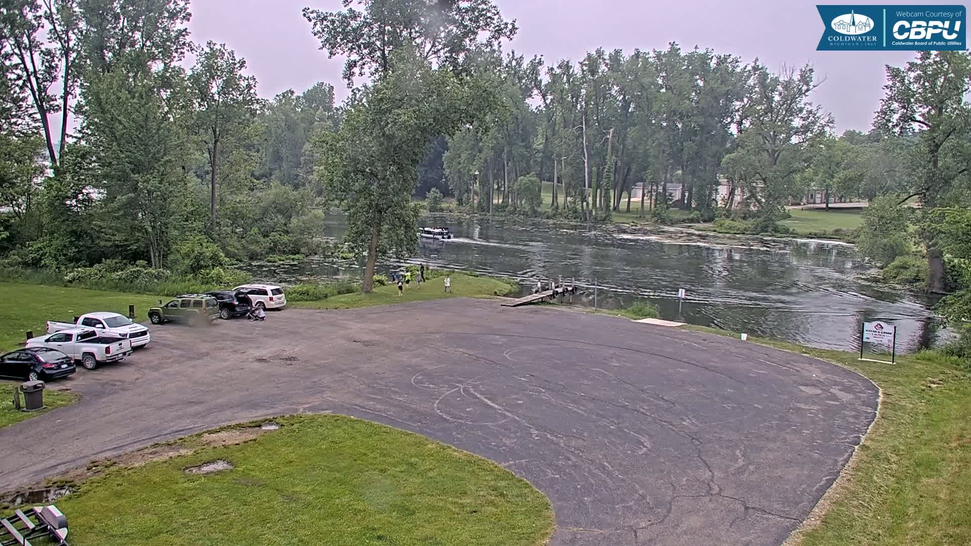 A paved boat launch area with several vehicles parked, overlooks a calm river surrounded by lush green trees under an overcast sky.
