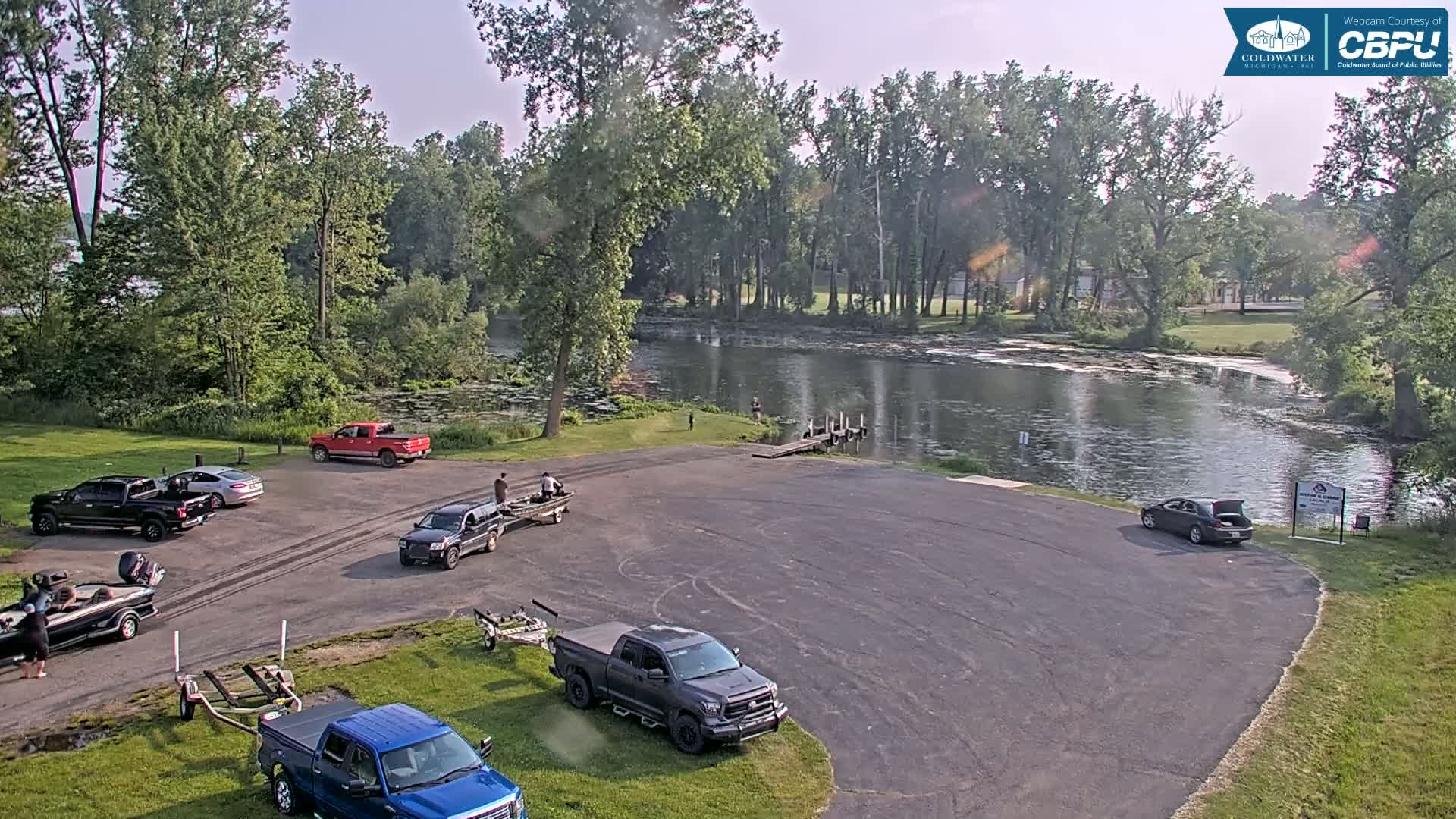 A paved boat launch area with several trucks and boats, surrounded by lush green trees, overlooks a calm river under a partly sunny sky.