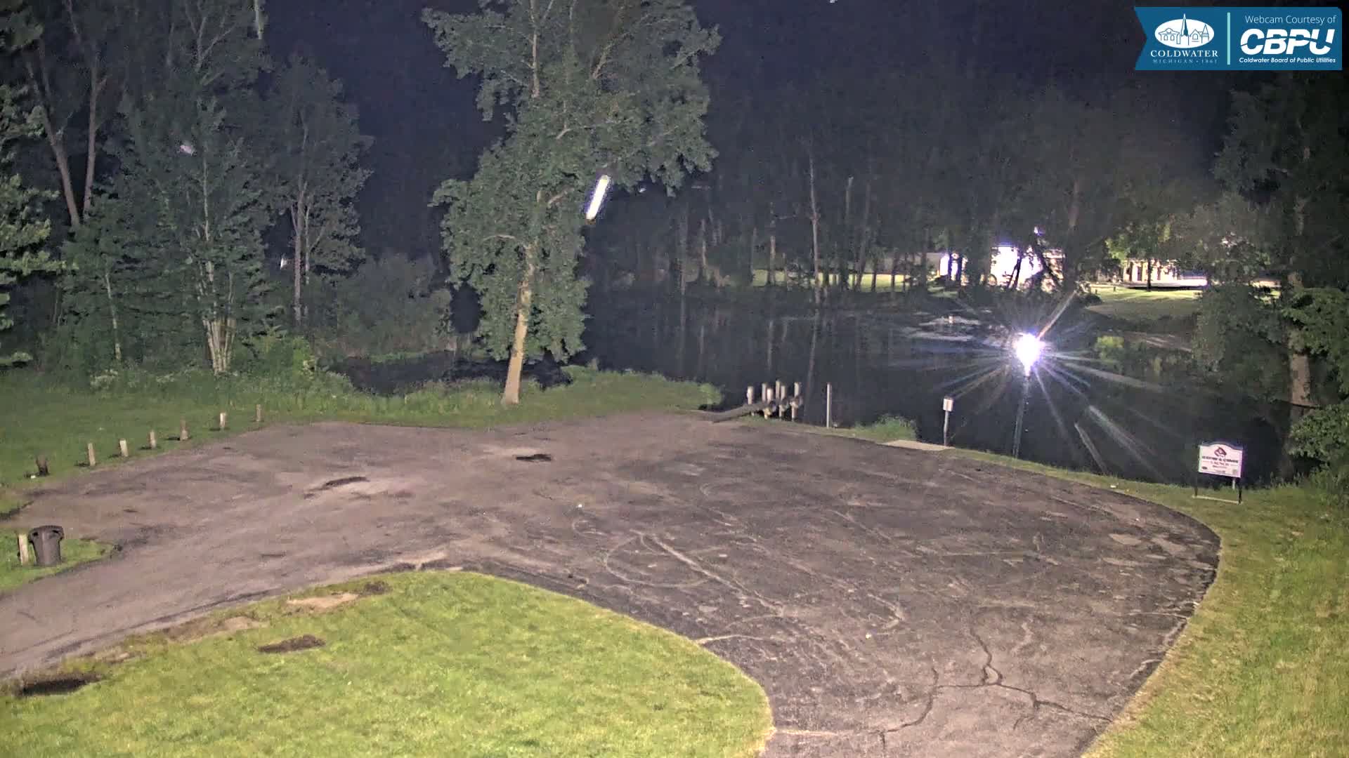 A nighttime view of an empty asphalt parking lot next to a dark body of water, surrounded by trees under a clear sky.