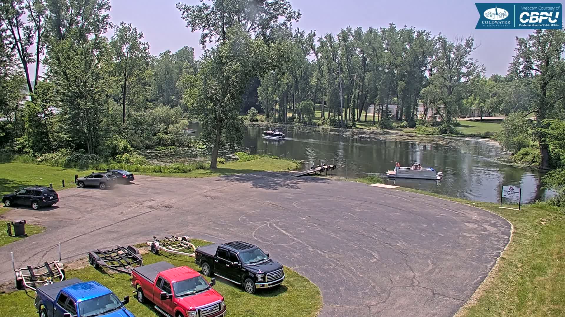 A parking lot with several vehicles and boat trailers overlooks a calm lake surrounded by lush green trees under a mostly sunny sky.