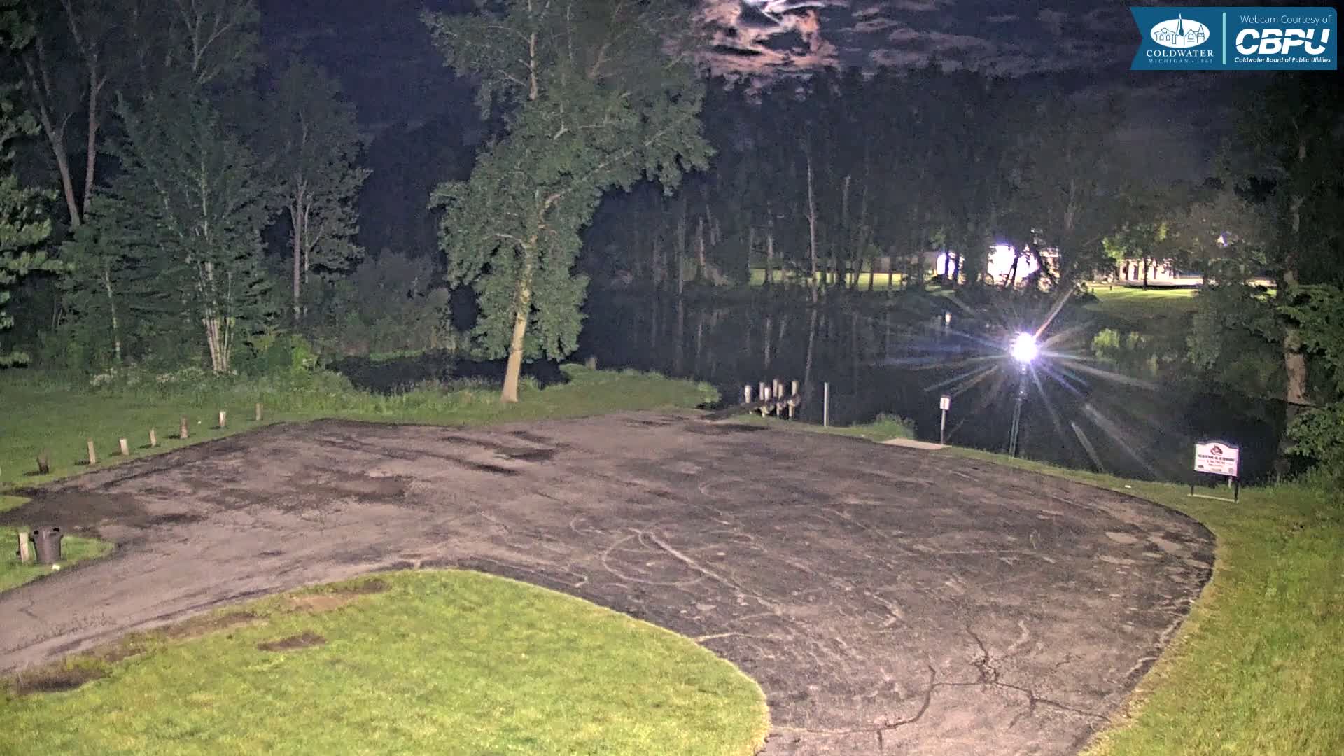 A nighttime view shows an empty asphalt parking lot next to a dark body of water, surrounded by trees under a partly cloudy sky.