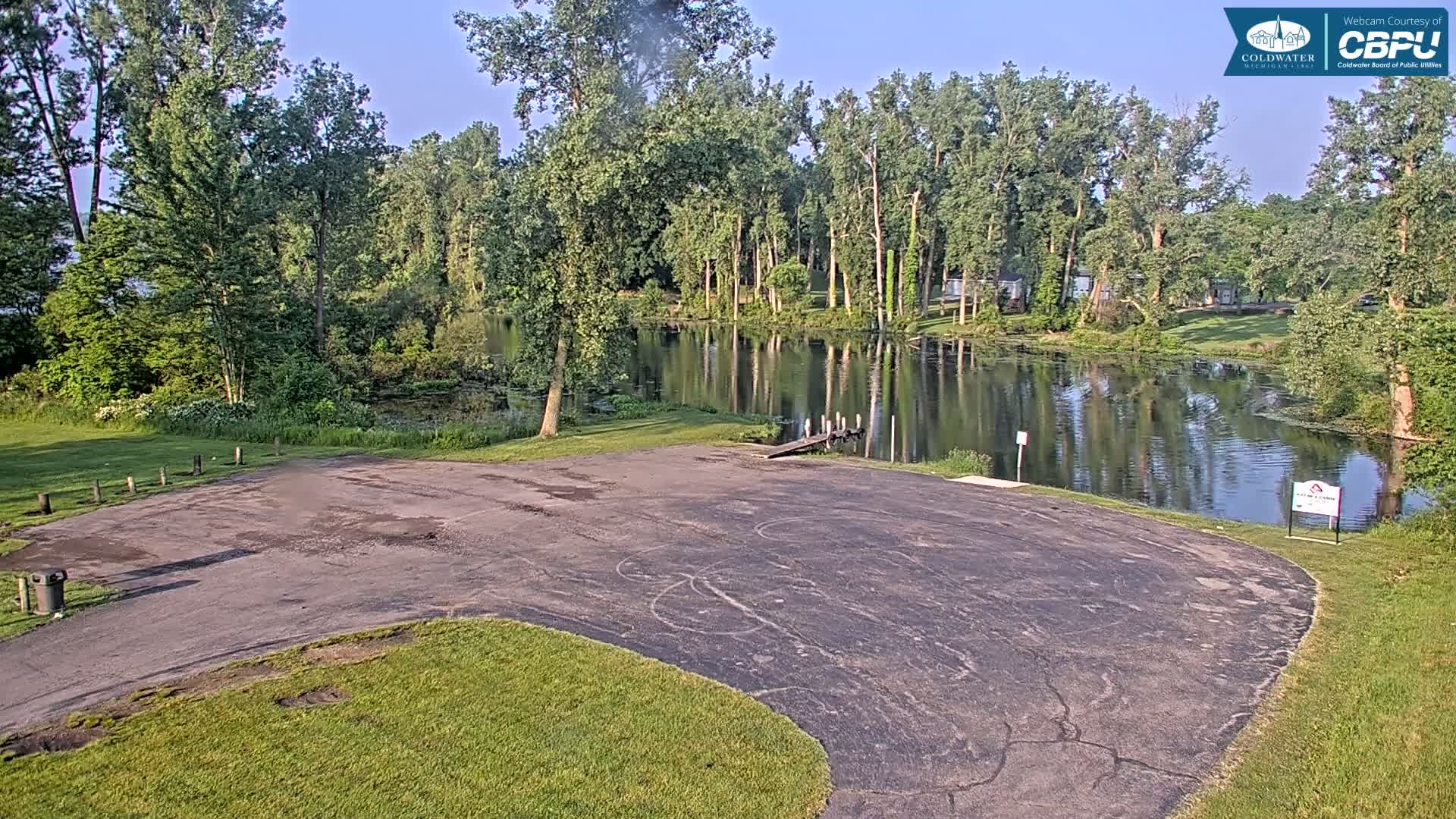 An empty asphalt boat launch area overlooks a calm, reflective lake surrounded by lush green trees on a sunny day.